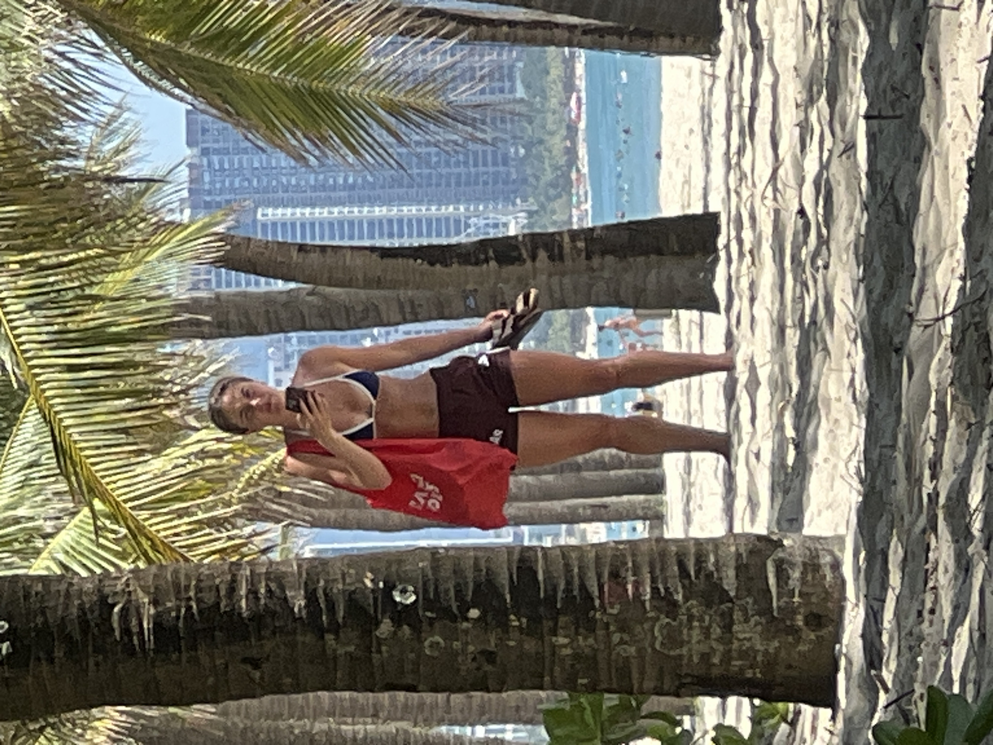 The image depicts a woman standing on a sandy beach in Da Nang, Vietnam, holding a red tote bag and a smartphone. She is wearing a dark blue and white bikini top and dark maroon shorts. Behind her, palm trees partially obscure a view of the ocean, with numerous beachgoers visible in the water and on the shore. In the background, the cityscape of Da Nang is prominent, featuring numerous tall buildings. The sand in the foreground is covered in shadows cast by the palm trees.