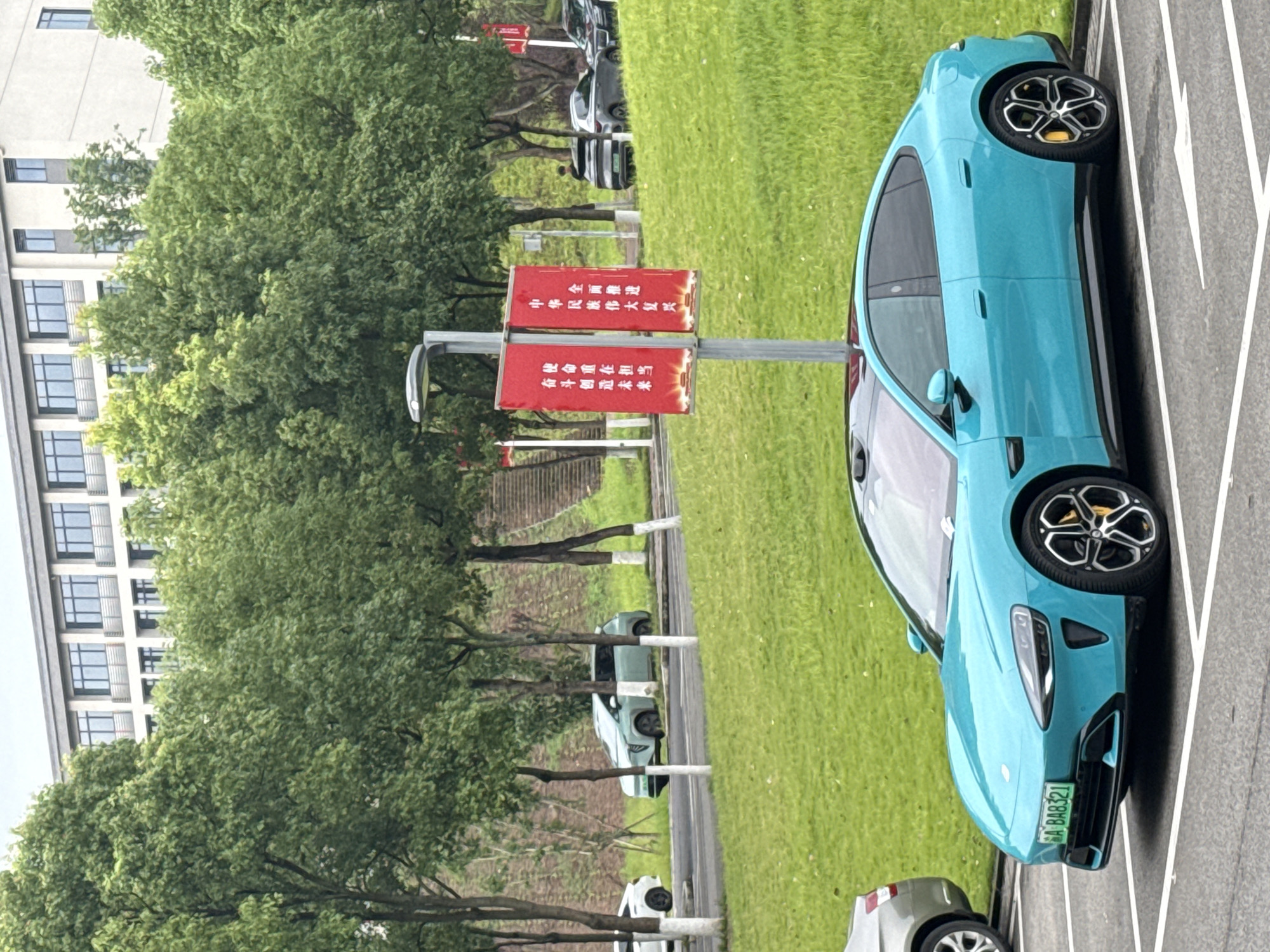 In Jiangjin, China, a turquoise electric sedan is parked in a lot on a grassy incline, adjacent to a road. In the background, a modern building with multiple windows stands behind a line of lush green trees. A lamppost with two red banners hangs in front of the trees. The license plate on the sedan reads "A BA8321." Other vehicles are visible on the road and in the distance. The sky is overcast.