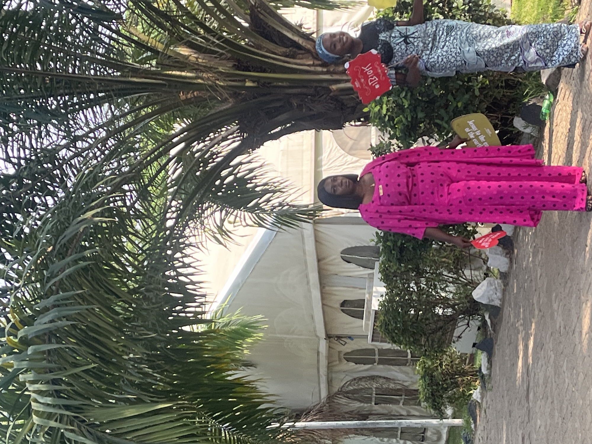 In Galadimawa Village, Nigeria, two women stand in front of a white tent. The woman closer to the viewer wears a bright pink, polka-dotted outfit and holds a red sign. Behind her and further away, the other woman wears a blue and gray patterned outfit, a blue headscarf, and also holds a red sign. A large palm tree dominates the left side of the image. The ground appears to be paved.