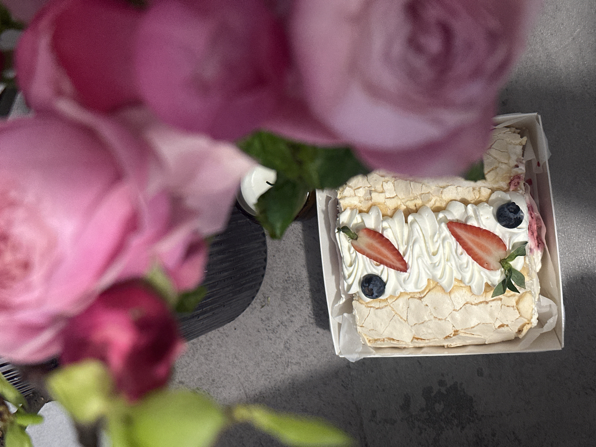 A high-angle shot depicts a cake in a white box placed on a gray surface. The cake is topped with white icing swirls, two sliced strawberries, and two blueberries. A floral arrangement with pink roses partially obscures the view of the cake. A small, cylindrical object resembling a candle is positioned next to the cake box. The setting is indoors, with diffused lighting. There is no evidence to suggest that the location is Kazan, Russia.