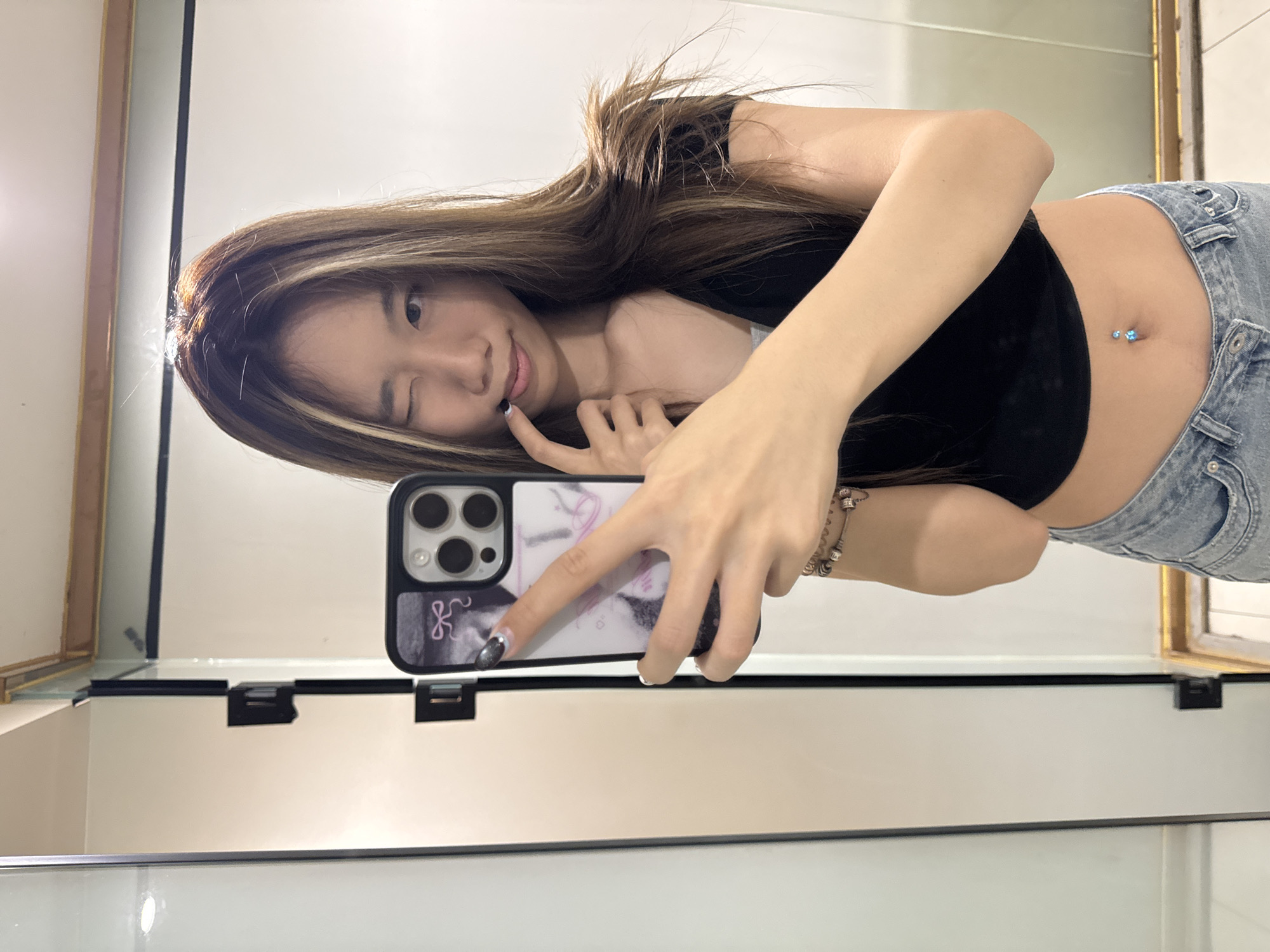 A young woman poses for a selfie in front of a bathroom mirror. She winks at the camera and holds her phone with a decorated case. The phone is a dark color with at least three lenses. She wears a black crop top and light-wash jeans. Her navel is pierced with what appears to be a blue barbell. The location in Yuzhong District, China is not discernible from the image.