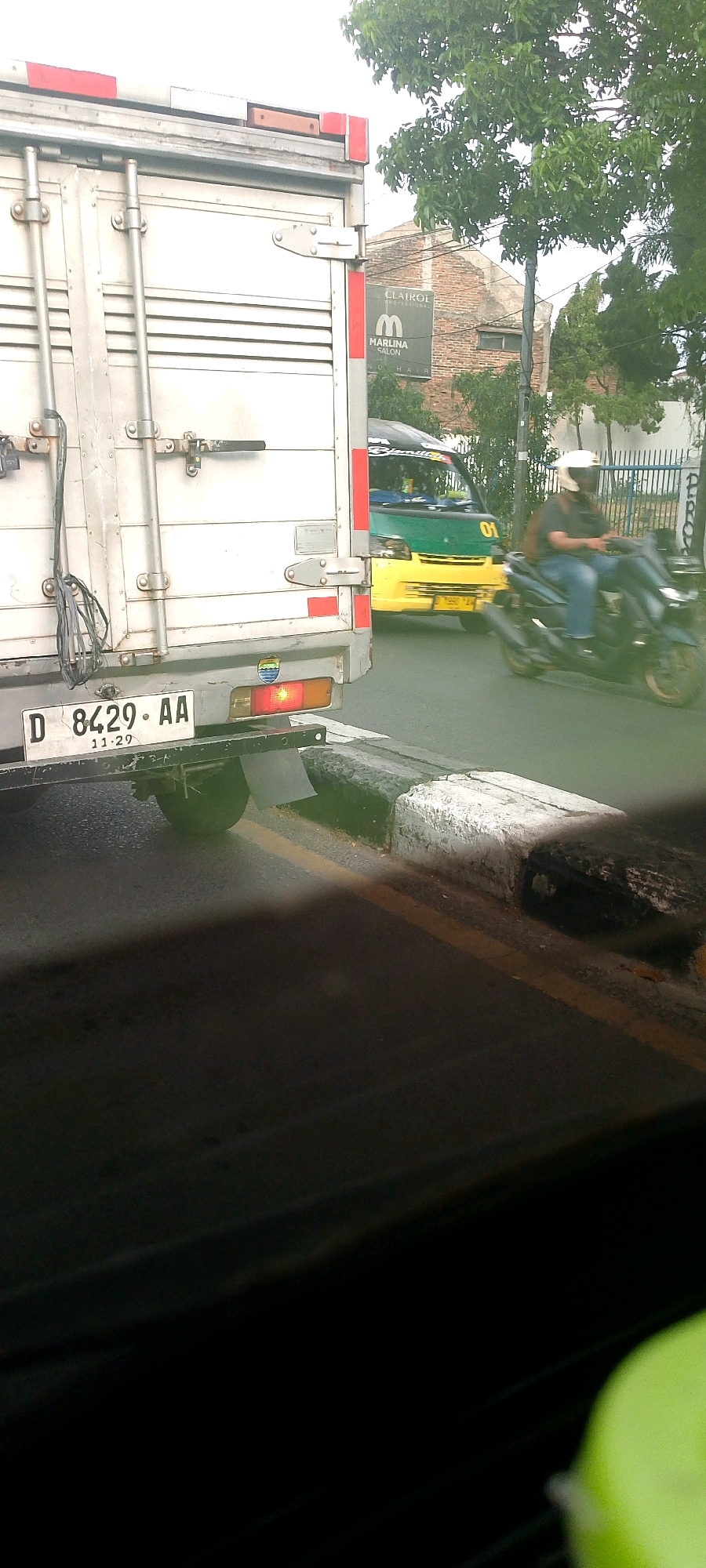 The image shows a street scene in Bandung City, Indonesia, with a focus on the rear of a white truck. The truck has a license plate that reads "D 8429 AA" with "11-29" below it. The truck's taillights are visible, with the right one illuminated. To the right of the truck, a yellow and green van is partially visible, and a person on a dark-colored motorcycle, wearing a white helmet and jeans, is riding past. In the background, a brick building with a sign that says "CLAIROL MARLINA SALON" is visible. Trees and a metal fence are also part of the environment. The road has a white and black curb with a yellow line marking the edge. The foreground is dark, suggesting the photo was taken from inside another vehicle.