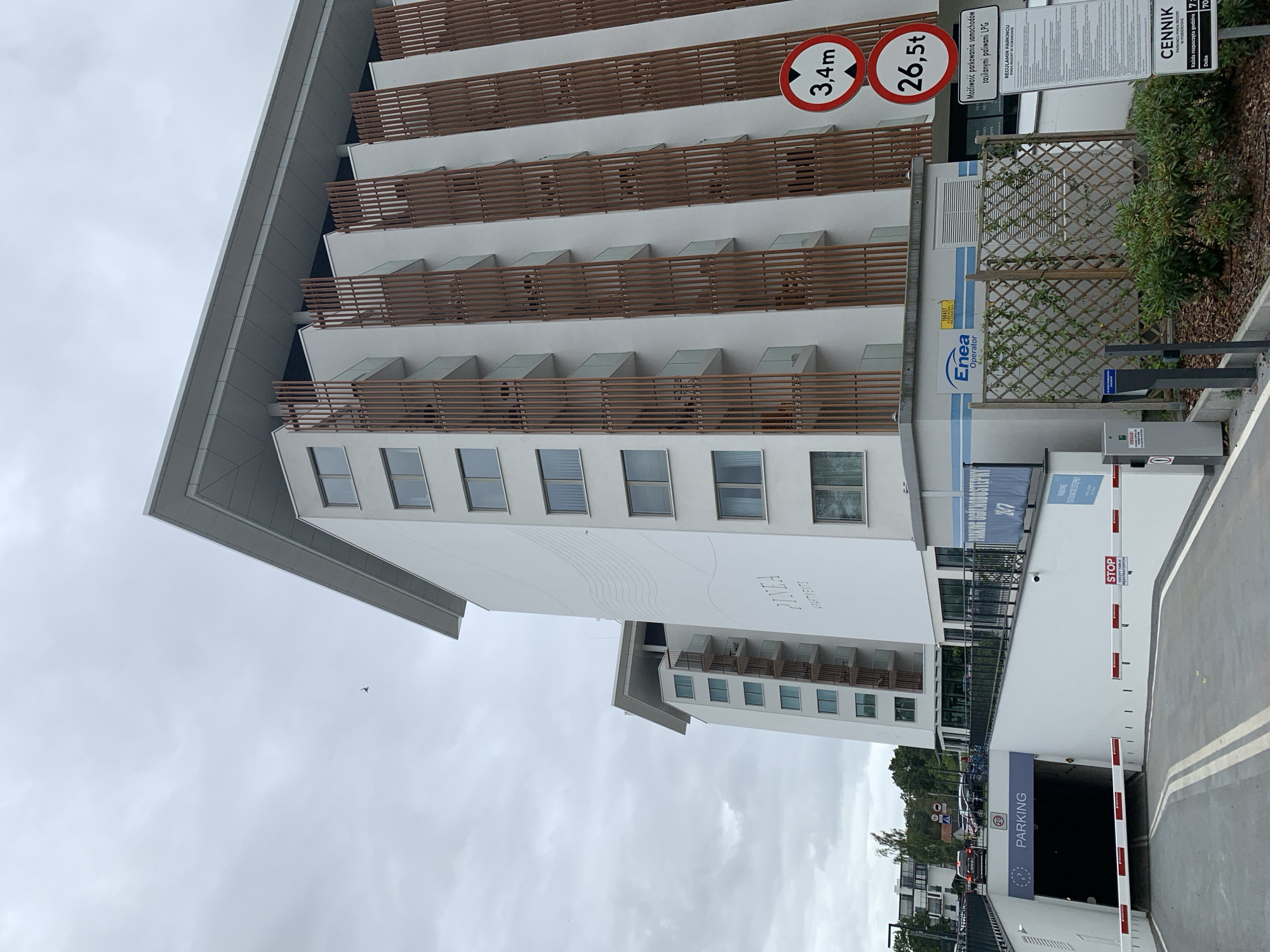 The image shows a modern apartment building in Pobierowo, Poland. The building is white with wooden vertical accents and balconies. In the foreground, there is an entrance to an underground parking garage. Traffic signs are visible, including one indicating a height restriction of 3.4 meters and another indicating a weight restriction of 26.5 tons. A sign for "PARKING" is above the entrance to the garage, and a blue banner on the side of the building reads "PARKING OGÓLNODOSTĘPNY 24/7". The sky is overcast. The building is labeled "PINEO APARTMENTS".