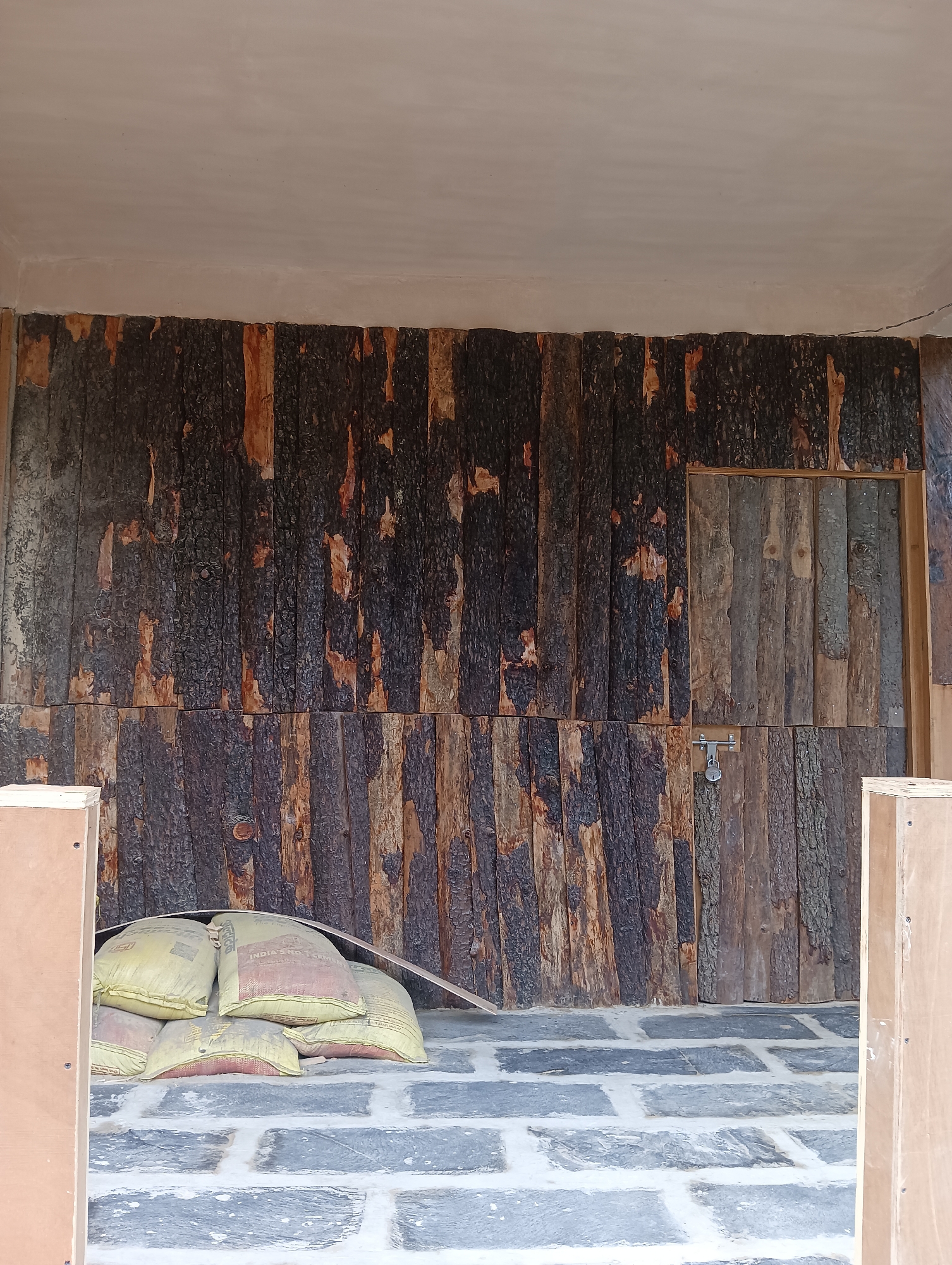 The image depicts a rustic exterior wall constructed from rough-hewn, dark-barked logs. The logs are vertically oriented and vary in thickness and color, with some displaying patches of lighter wood. A section of the wall, to the right, is covered with smaller, more uniformly sized vertical wooden planks. A metal latch and padlock are affixed to this planked section, suggesting a door or a locked compartment. In the foreground, a pile of several large, yellow sacks, likely containing cement or building materials, rests on a stone-tiled floor. The floor is composed of large, irregularly shaped grey stones with white grout. The scene is framed by unfinished wooden structures on the left and right edges of the image, implying ongoing construction or an undeveloped area. The overall impression is one of a rural or construction site environment, possibly in the city of Goshal, India.