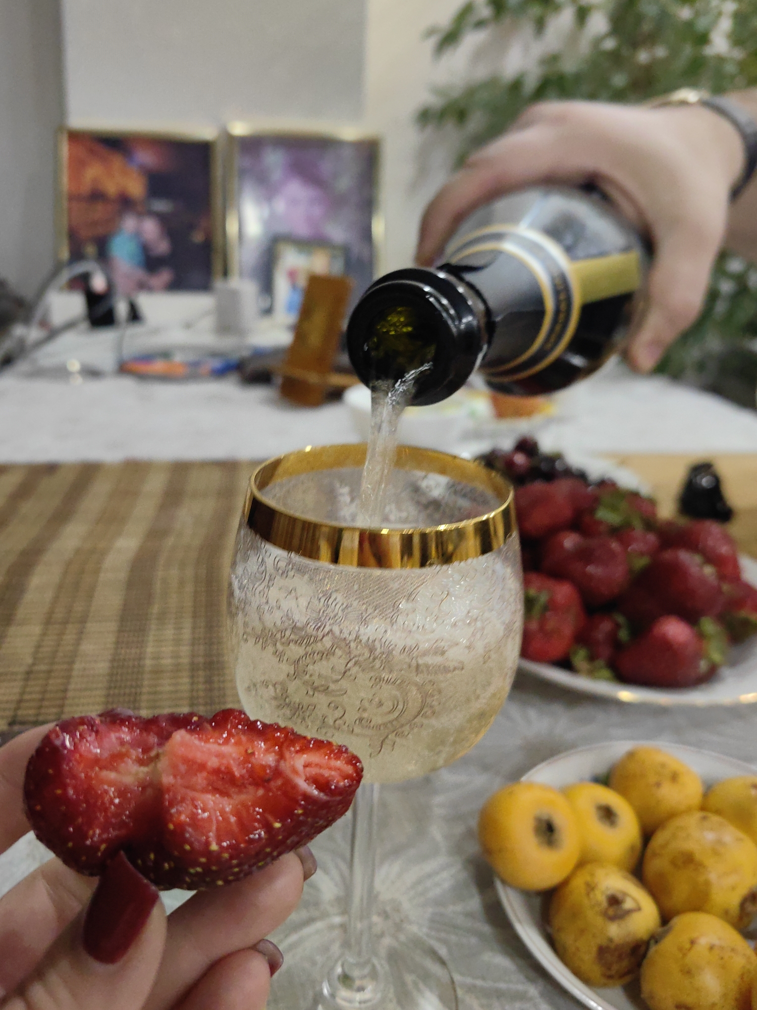 In a close-up shot, a person's hand pours a golden liquid from a dark bottle into a crystal glass with a gold rim. The glass is adorned with an intricate etched pattern and is already partially filled, with tiny bubbles rising to the surface. In the foreground, a halved strawberry, showcasing its vibrant red flesh and seeds, is held delicately between fingertips with dark red nail polish. In the background, a plate overflows with ripe strawberries and some darker berries, while another plate holds small, round yellow fruits. The scene is set on a table covered with a light-colored tablecloth patterned with a delicate floral design. Two framed pictures hang on the wall in the soft-focus background, one appearing to depict people in what looks like a city environment. The overall impression is one of a casual, festive gathering or a moment of personal indulgence. The specific location is not identifiable from the visual cues.