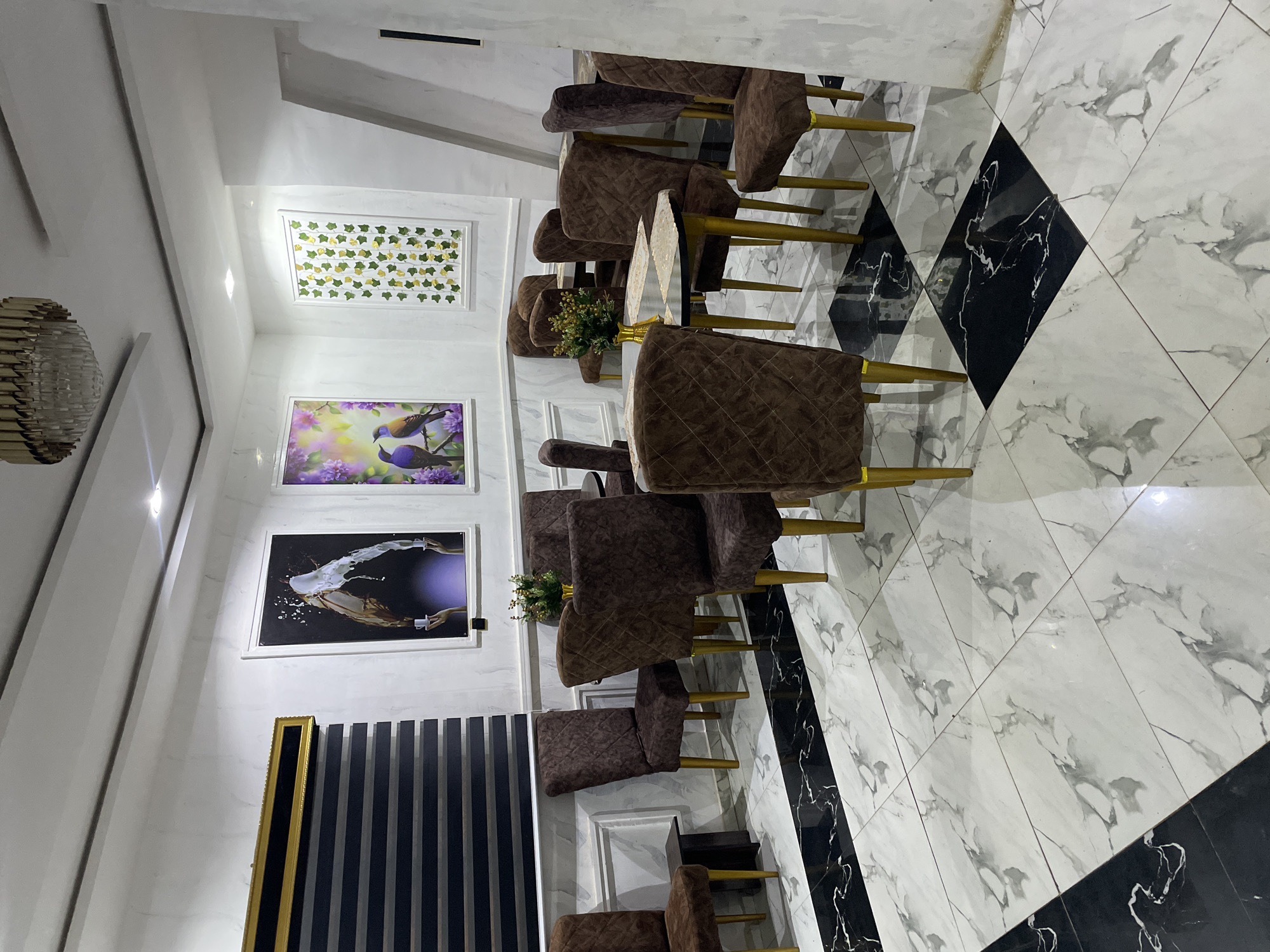 The image shows an indoor dining area, likely in a residence or small establishment in Tumfure, Nigeria. The focal point is a set of upholstered brown chairs with gold-colored legs arranged around a table. The floor is tiled with a white marble pattern interspersed with black diamond-shaped tiles. Three framed pictures decorate the wall, along with a set of vertical striped black panels. The image is angled, providing a skewed perspective of the room.