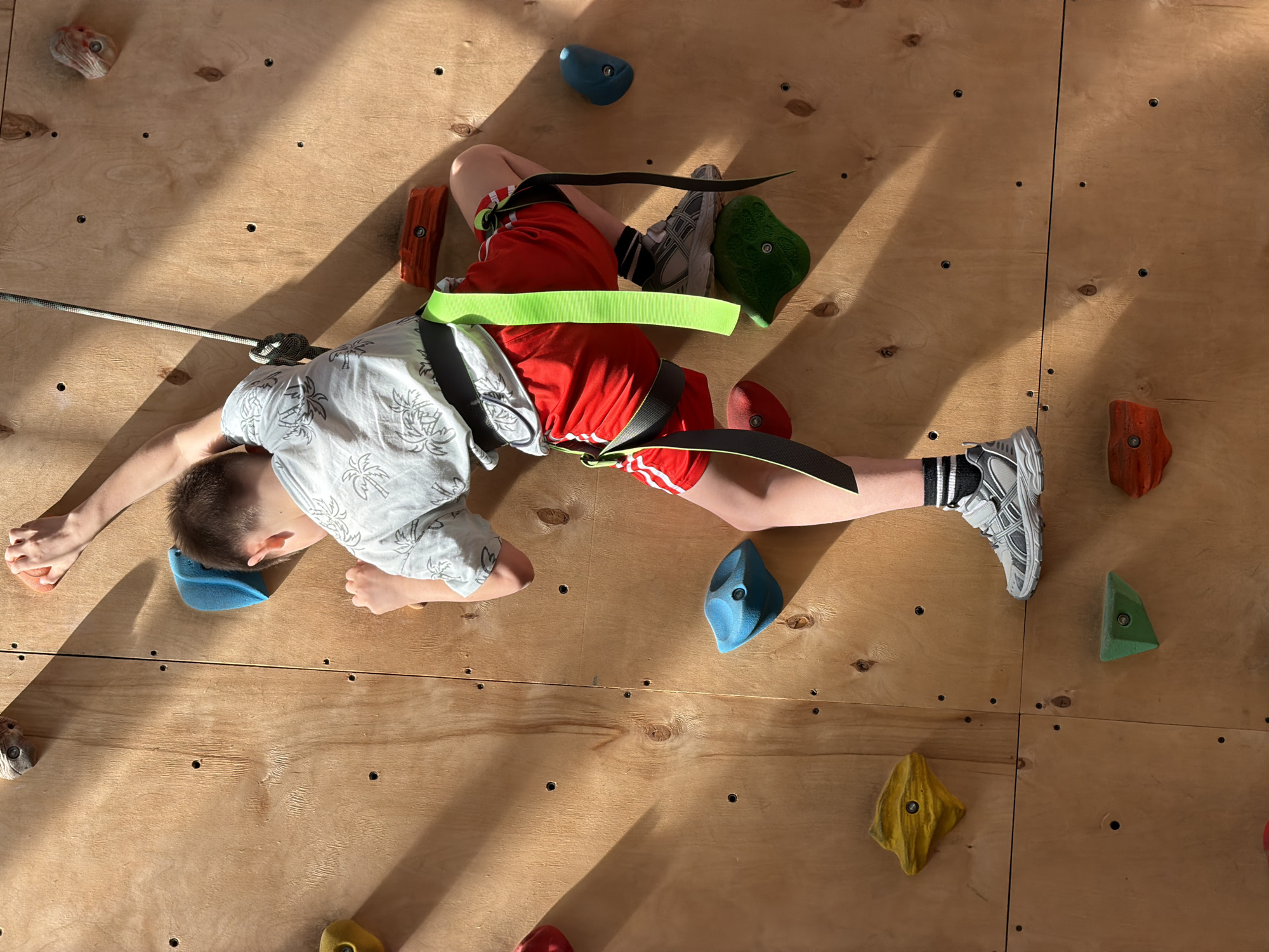 A young person, wearing a patterned white short-sleeved shirt and red shorts with white stripes, is suspended on a climbing wall. The individual is secured by a climbing harness with neon green straps and is connected to a rope. The climbing wall is constructed of plywood and features various colored climbing holds. The location is the city of Izhevsk, Russia.