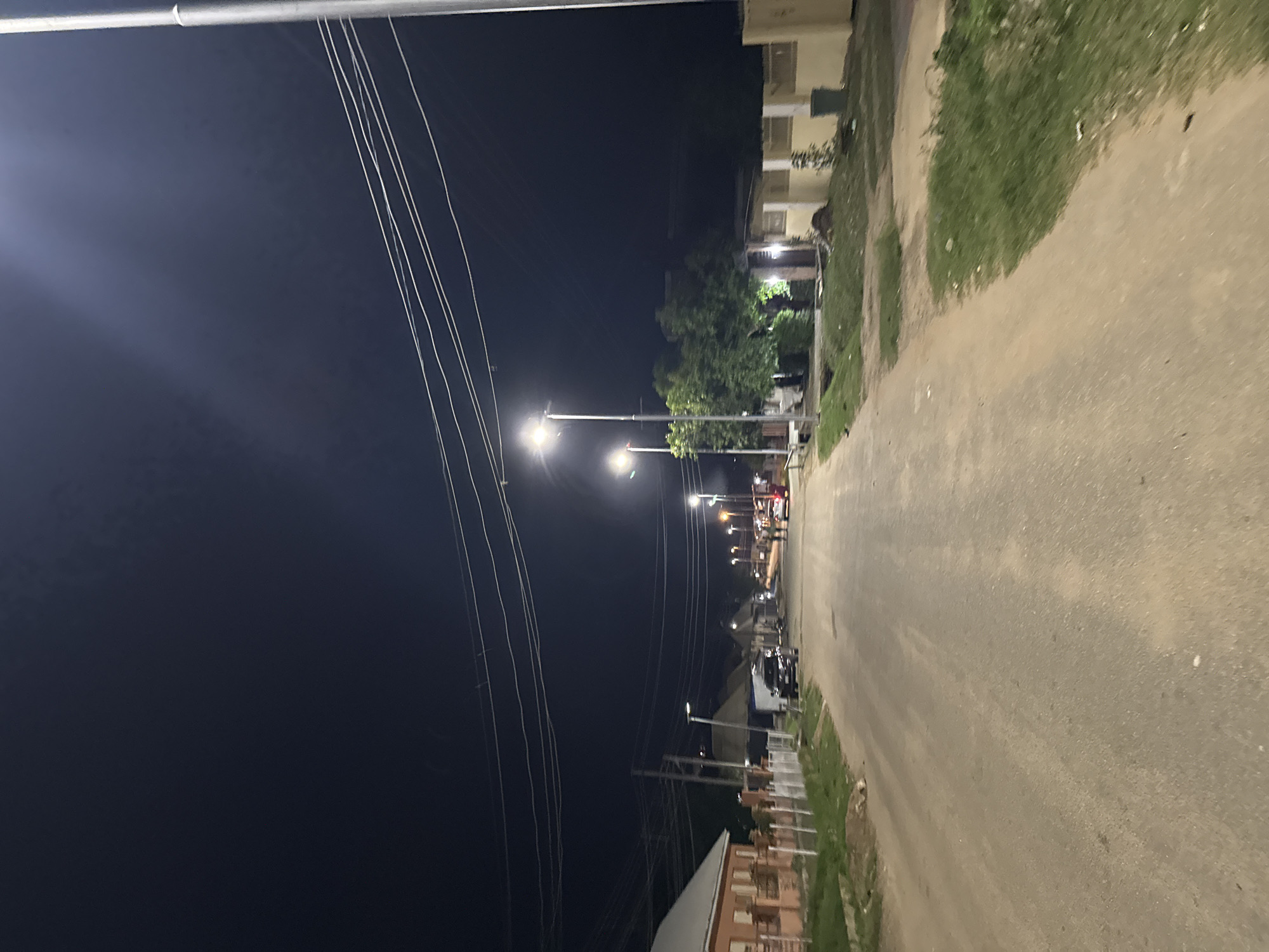 A street scene at night in Abuja, Nigeria, depicts a paved road illuminated by streetlights. The road is flanked by buildings and vegetation. Several streetlights are visible, casting pools of light onto the road and the surrounding area. On the left side of the road, a residential building with an orange facade and white-trimmed windows is visible, bordered by a brick wall and fencing. Further down the road, more buildings and trees can be seen. A dark-colored SUV is parked on the side of the road. A few individuals are walking on the road in the distance, and a red vehicle is also present. The sky is dark, indicating nighttime. Power lines are strung across the upper portion of the image. The overall scene suggests a residential or commercial street in a developing urban area.