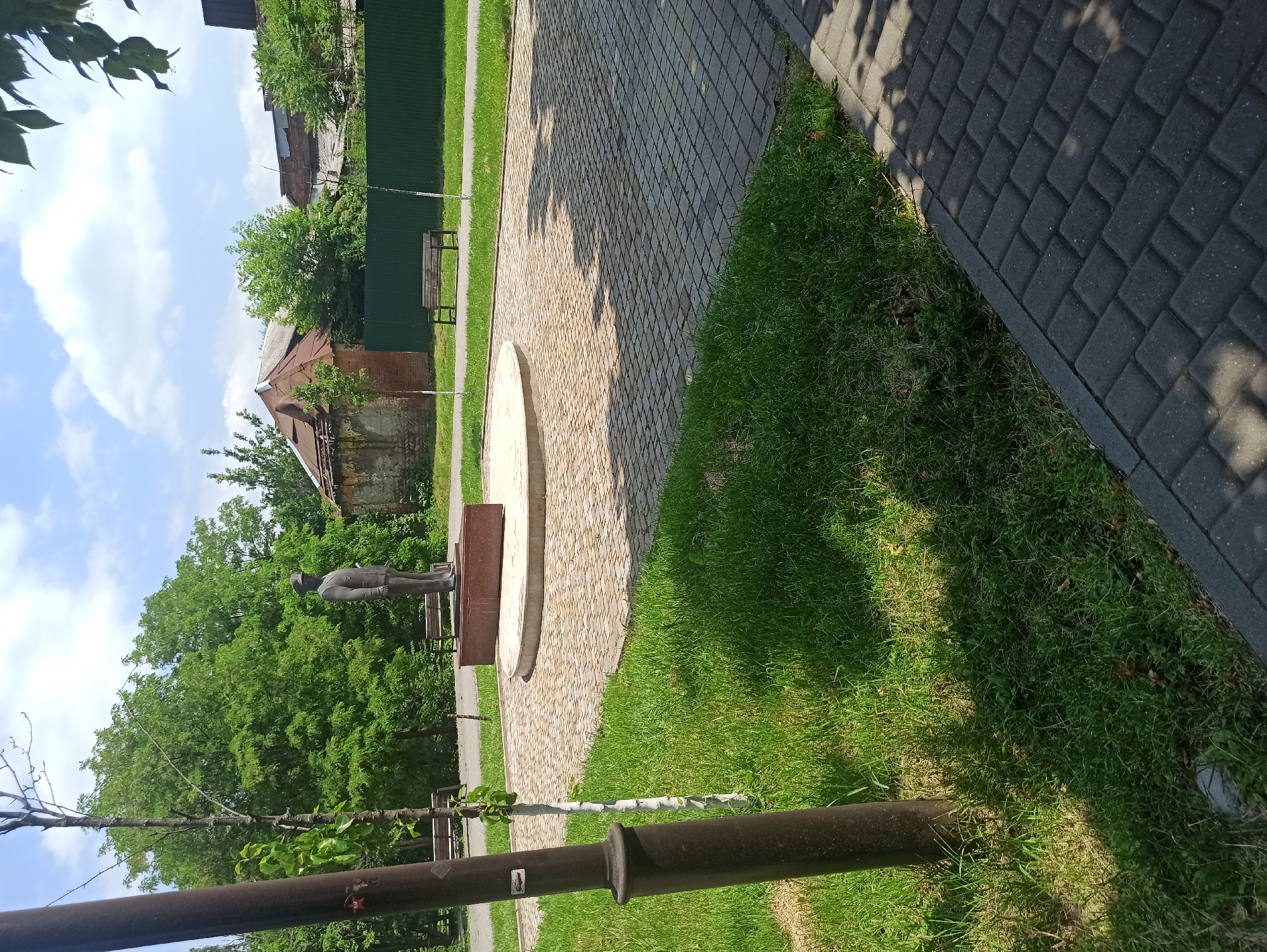 A statue of a man in a uniform stands on a circular, paved platform in a park. He is facing to the left of the frame. In the background, there are trees, a green fence, and buildings with dilapidated roofs. Two wooden benches are visible on the left and right sides of the park. The ground is covered with grass and paved with stone tiles. The sky is blue with scattered clouds. A dark pole stands in the foreground on the left side of the image.