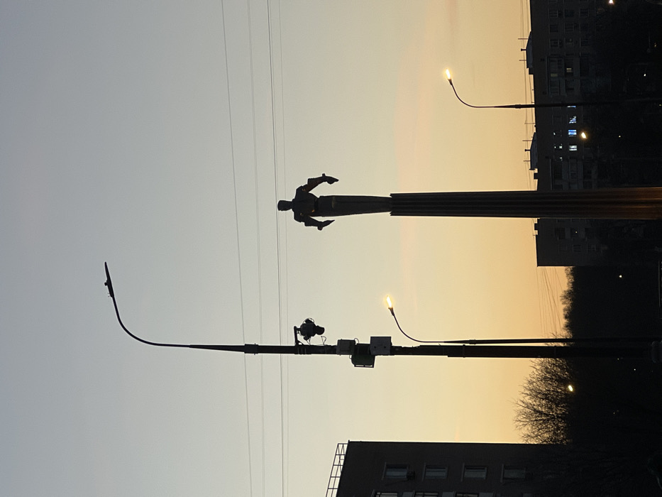 The image captures an urban street scene during twilight, characterized by artificial illumination against a gradient sky transitioning from pale blue overhead to warm orange-yellow near the horizon.
Dominating the central-right is the Monument to the Conquerors of Space, located in Moscow, Russia, near the VDNKh complex. The monument consists of a tall, fluted column, dark in silhouette, topped by a stylized, silhouetted male figure with arms extended sideways.
To the left of the monument stands a tall, dark streetlamp with a curved arm holding a single overhead light fixture. Midway down its pole, a downward-facing surveillance camera or similar sensor and multiple utility boxes are attached. A shorter, illuminated streetlamp with a yellow-orange glow is positioned slightly in front and to the right of the tall lamp. A third, similarly illuminated streetlamp is visible on the far right.
In the background, several multi-story buildings are discernible, mostly silhouetted against the sky. A building on the far left shows visible windows. Further right, another building has several illuminated windows on its lower floors. A line of dormant trees or bushes forms a dark band along the middle ground. Multiple thin, dark utility lines span horizontally across the upper portion of the sky. No human figures are overtly visible.