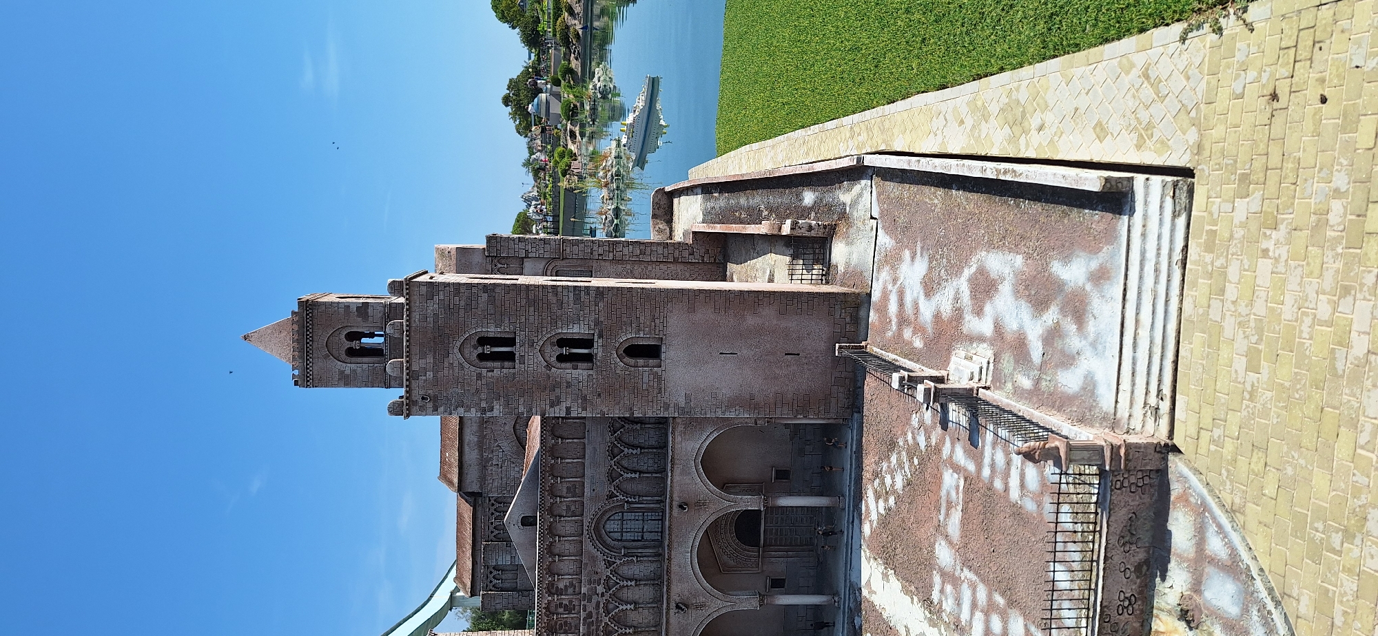 The image shows a miniature model of the Duomo di Cefalù, a cathedral located in Cefalù, Sicily, Italy. The model is situated in an outdoor setting with a clear blue sky. In the background, there is a body of water with a miniature boat and other miniature buildings and greenery, possibly representing a theme park or model village. The Duomo model features a prominent bell tower with arched windows and a pyramidal roof, and the main body of the cathedral has intricate architectural details like arches and columns. A paved courtyard with some patchy discoloration is in front of the cathedral, bordered by a fence. A strip of bright green grass is visible to the right of the model, adjacent to a brick pathway. There are no people visible in the image, only small figures that appear to be part of the model itself. The scene is brightly lit by sunlight, casting shadows. The specific location in Rimini, Italy, is not identifiable from the image itself; it's a model of a building from Sicily.