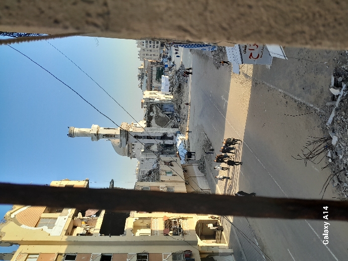 The image displays a street scene in Khan Yunis, Palestinian Territory, captured from an elevated perspective, possibly through a window frame, with a dark vertical beam partially obstructing the left-center view. The environment is characterized by significant urban damage under a clear blue sky.
In the center-background, a white mosque with a prominent minaret and dome stands, appearing structurally compromised with visible damage to its facade. To the left of the mosque, a multi-story building exhibits extensive structural damage, including large sections of missing walls, exposed interior rooms, and debris hanging from balconies. Other buildings in the background also show varying degrees of damage.
The street itself is wide, appearing unpaved or covered in dirt and rubble, particularly in the foreground and along the edges. Piles of concrete and other debris are visible along the roadside.
Multiple individuals are present on the street. A group of approximately ten individuals, predominantly men in dark attire, walk in a loose formation on the right side of the road, moving away from the viewer. Smaller groups and solitary figures are also visible further down the street, near the mosque, and along the sidewalk to the left, some appearing to be near the entrance of the damaged building. In the far background on the right, a cluster of white tent-like structures is discernible. A partial white sign with red Arabic script is visible on the right side of the street, near the foreground. Electrical wires traverse the sky above the street.