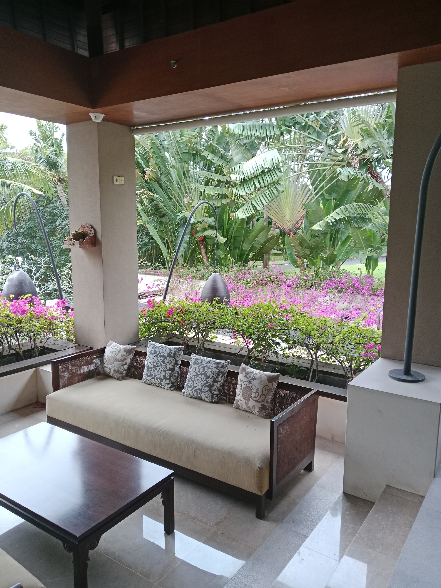 A covered patio area in Kuta Selatan, Indonesia features a tan sofa with decorative pillows and a dark wood coffee table on a tiled floor. Beyond the patio, a lush garden with vibrant pink flowers and large tropical plants is visible. Two large, dark, bell-shaped hanging objects are positioned on either side of the patio opening. A security camera is mounted on the ceiling of the patio.