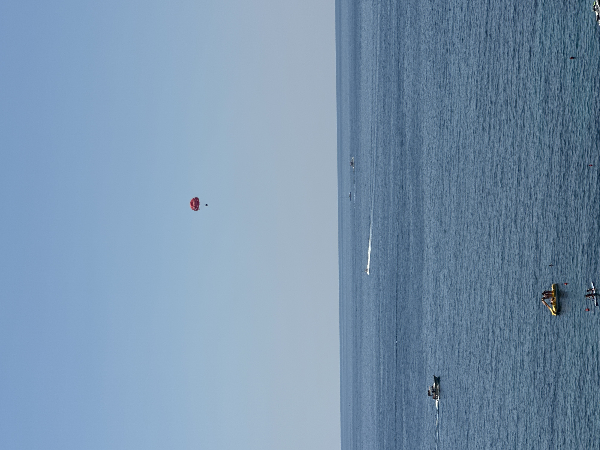 This image depicts a clear, sunny day over the Adriatic Sea near Kamenovo, Montenegro. The horizon is sharply defined, separating the deep blue water from a pale blue sky.
In the sky, a single red parasail with a person attached is visible in the upper center of the frame.
The sea is populated with various watercraft. Towards the left side of the image, a small white motorboat is moving, leaving a white wake. Further out, a smaller boat is barely visible. In the lower part of the frame, a yellow pedal boat with at least two people on it is present. Below that, a paddleboard with two individuals is also visible. There are also small red buoys scattered in the water.