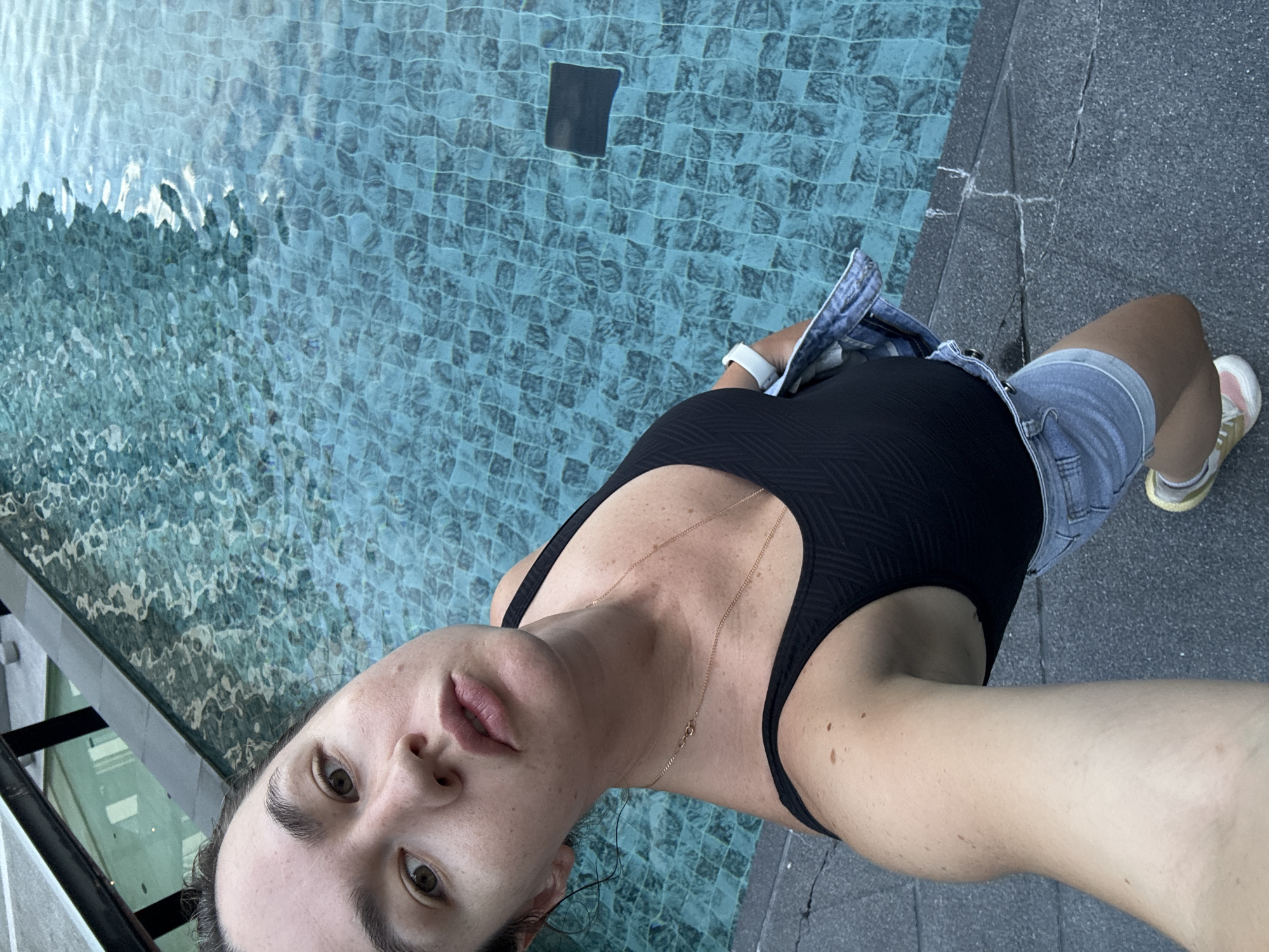 A woman stands on the edge of a tiled swimming pool in Ratsada, Thailand. She is wearing a black swimsuit, denim shorts, and a denim jacket draped over her shoulder. The woman is looking up at the camera with a neutral expression. The pool water is blue and reflects the light. The background shows a glimpse of the environment, possibly a hotel or resort.
