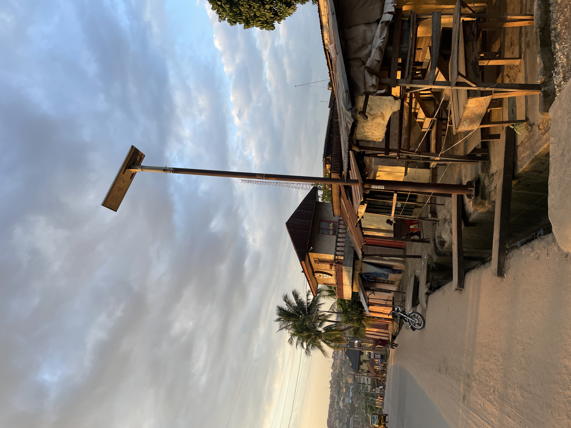 This is a street-level, eye-level, full shot captured in Nkpor, Nigeria, likely during a warm and possibly humid evening given the golden hour lighting and presence of clouds. The scene depicts a street with a dirt shoulder on the left, leading into a paved road. Along the right side of the road, a row of simple, open-air structures with corrugated metal roofs, made of wood and possibly tin, are present. These structures appear to be informal shops or market stalls.
In the foreground, a large, open drainage ditch runs alongside the stalls, with wooden planks placed across it in several sections, serving as makeshift bridges. A solitary motorcycle is parked on the dirt shoulder. Further down the street, a yellow auto-rickshaw is visible, along with a few pedestrians.
Dominating the mid-ground is a tall, slender pole topped with a solar panel, indicating a form of off-grid street lighting. Behind the stalls, a two-story building with a red roof and balconies stands out, flanked by palm trees. In the background, a sprawling cityscape with numerous buildings is visible, stretching up a gentle incline. The sky above is filled with dramatic, textured clouds, some appearing heavy and dark, others illuminated by the setting sun, creating a dynamic contrast of light and shadow across the scene. The overall impression is one of a busy, functional urban environment in a developing country, characterized by informal economies and resourceful infrastructure.