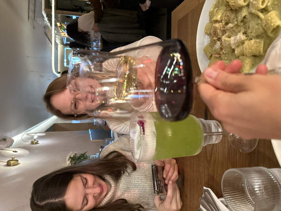 Two young women are at a restaurant. One woman is seen through two wine glasses, and another woman is looking at her phone with a green drink on the table. A partially visible plate of pasta sits to the right, and cutlery is neatly placed. The background shows restaurant lighting fixtures and other patrons.