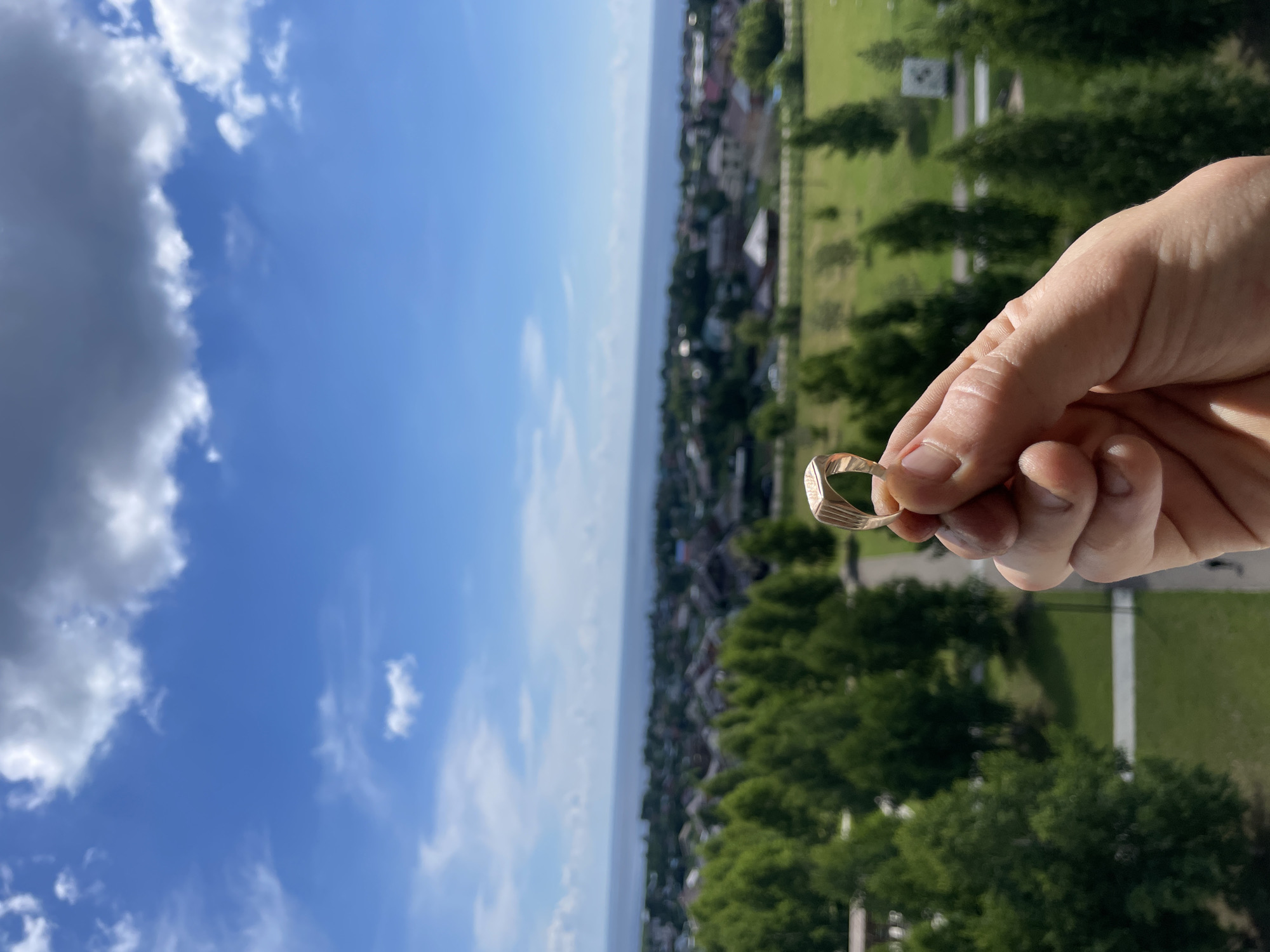 A hand holds a gold signet ring in the foreground, with a blurred cityscape and a body of water in the background. The cityscape appears to be a residential area with houses and trees. A body of water is visible on the horizon, possibly a lake or the sea. The sky is blue with scattered clouds. The overall scene suggests a sunny day with a picturesque view. The image was taken in Laishevo, Russia.