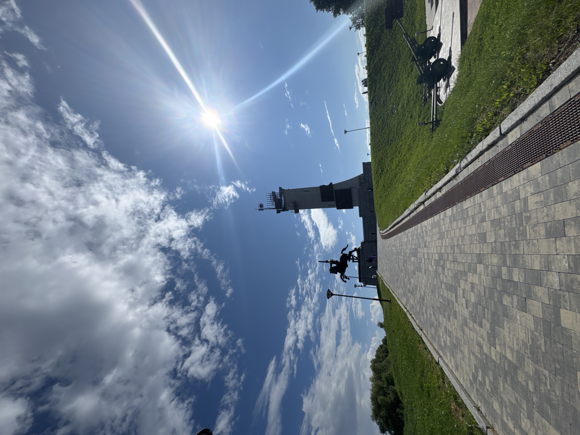 In Veliky Novgorod, Russia, a wide, paved walkway leads uphill towards a monument featuring a statue of a man on horseback. To the right of the walkway, two cannons are displayed on a grassy slope. In the distance, a tall, slender tower stands against a bright blue sky dotted with white clouds. The sun is visible, casting a strong glare. Several people are seen near the monument and on the grassy hill.