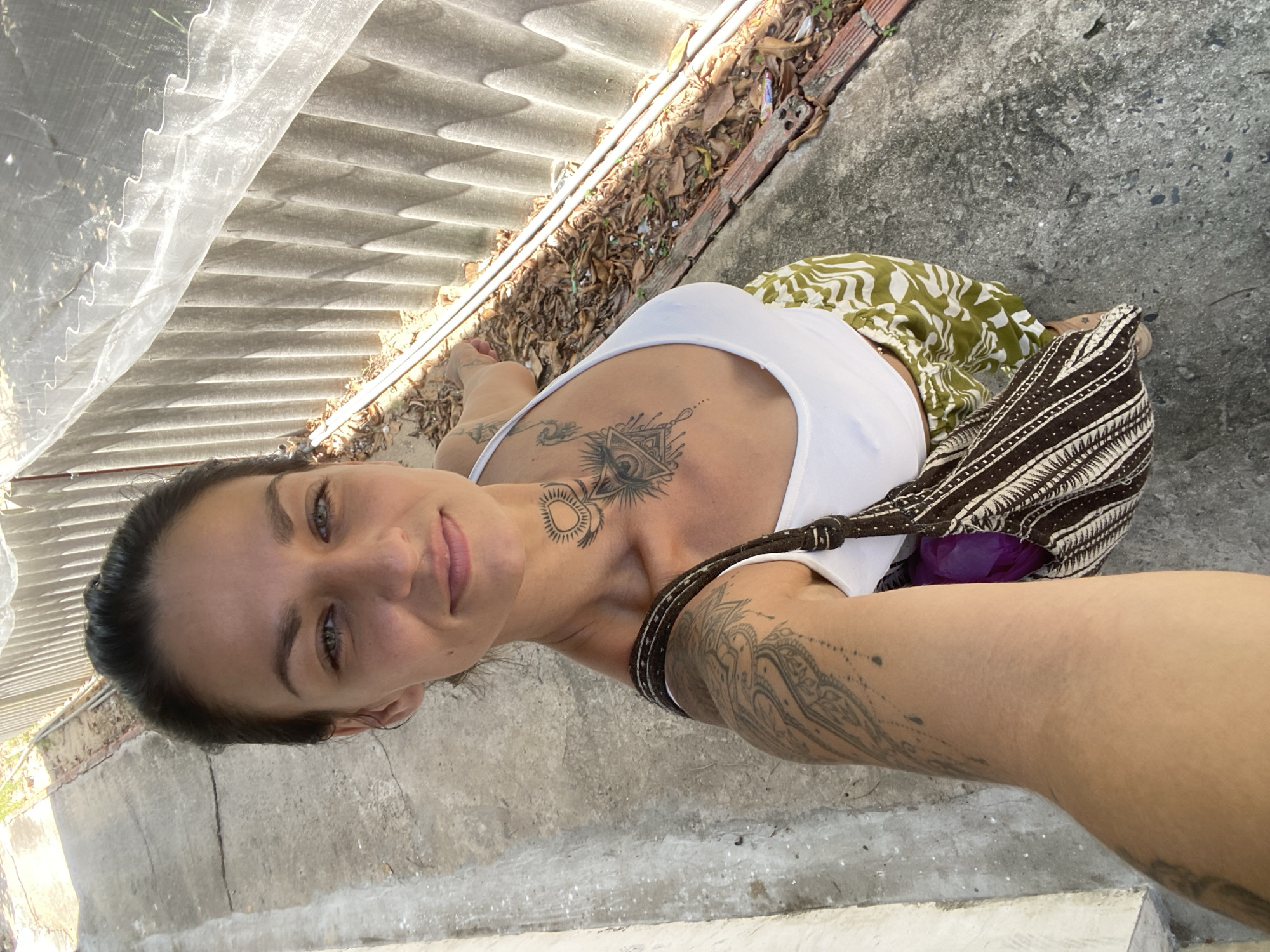 A woman with dark hair pulled back and green eyes is taking a selfie outdoors. She is wearing a white tank top, patterned green and white pants, and a woven shoulder bag. She has tattoos on her chest, neck, and left arm. The background appears to be a concrete path next to a corrugated metal fence, with dry leaves and debris on the ground. The lighting suggests it is daytime and sunny. The location is identified as Mũi Né, Vietnam.