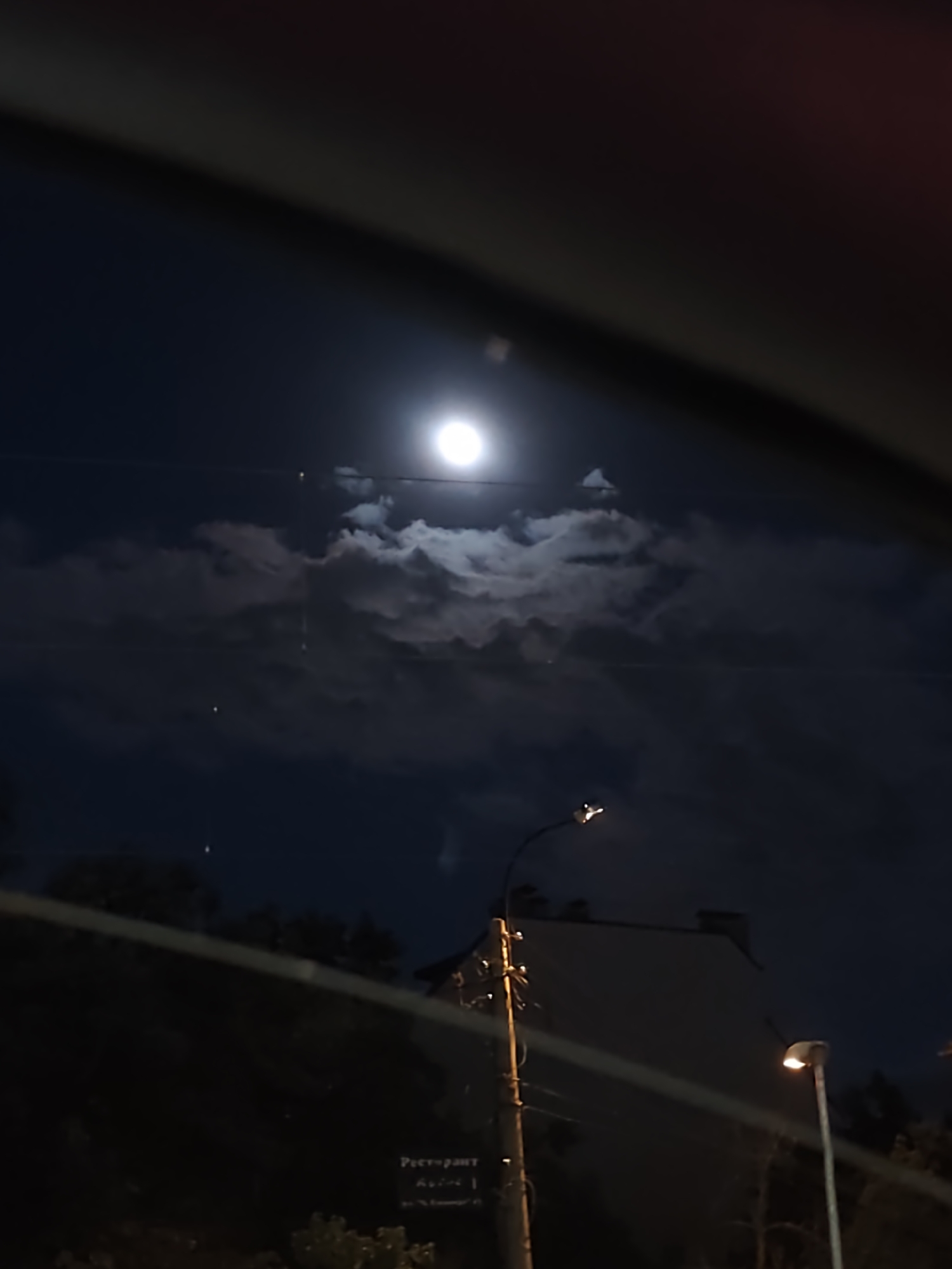 Night scene observed through a vehicle window featuring a full moon partially obscured by clouds in a dark blue sky. Below the sky, silhouetted trees are visible, and a light-colored building with a slightly peaked roof can be seen. Two illuminated streetlights are also visible in the foreground. A wooden pole with electrical wires is situated near the building. A sign with the word "PECTOPANT" (restaurant in Bulgarian) can be seen, suggesting the location might be in Bulgaria. Given the architecture and available information, the location is likely the city of Sofia, Bulgaria. No people are visible.