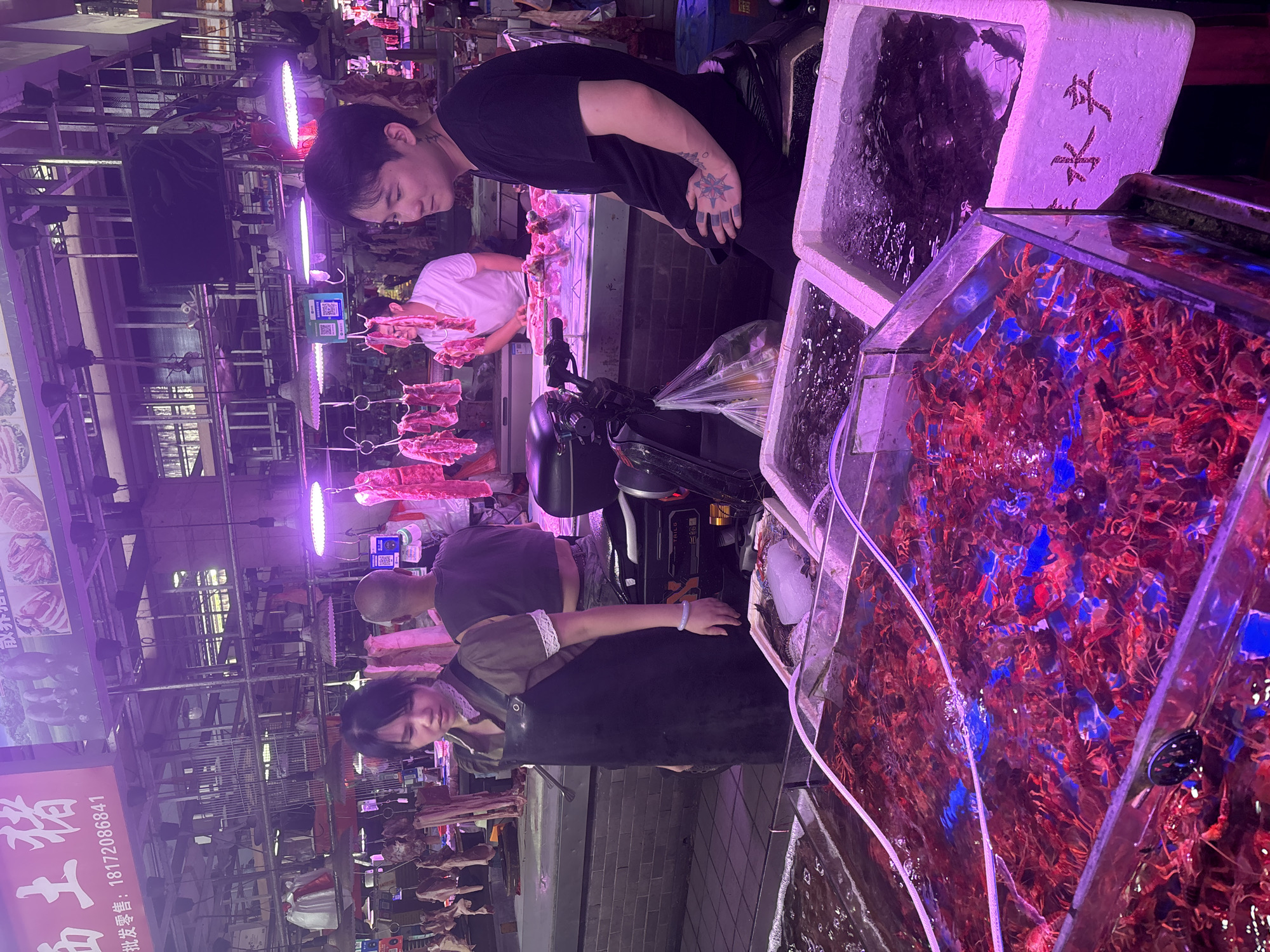 This is an indoor market scene in Nanhai District, China. In the foreground, a large rectangular glass tank is filled with live red crawfish swimming in water, illuminated by blue and purple lights. To the right of the crawfish tank, two white styrofoam boxes are filled with dark-colored seafood, one with bubbles indicating aeration.
In the middle ground, a woman wearing a dark apron over a light-colored shirt with a lace collar stands behind a counter, looking down. To her right, a bald man wearing a dark t-shirt is also standing. Behind them, a man in a white t-shirt is visible, appearing to be working at a meat stall where various cuts of pork are hanging from hooks.
In the background, more meat products are displayed under bright purple lighting. A scooter with a plastic bag hanging from its handlebars is parked to the right of the woman. The overall environment suggests a bustling food market, likely specializing in fresh seafood and meat. Above the stalls, overhead lights and signage are visible.