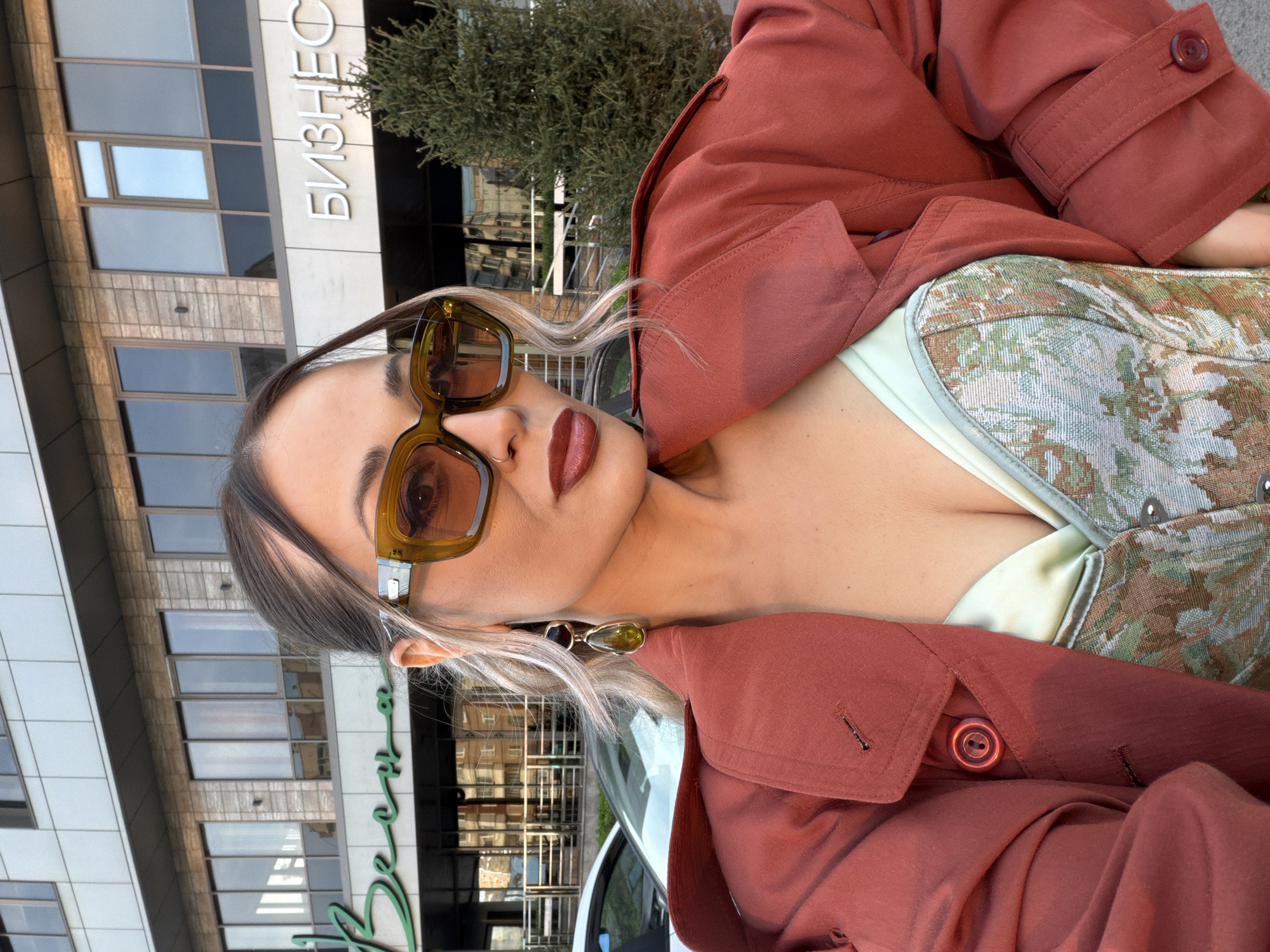 A woman with fair skin, brown-grey hair, and lipstick poses in front of a building in Krasnoyarsk, Russia. The building has the word “БИЗНЕС” on it. She is wearing a rust-colored jacket and a floral corset. She is also wearing sunglasses and earrings.