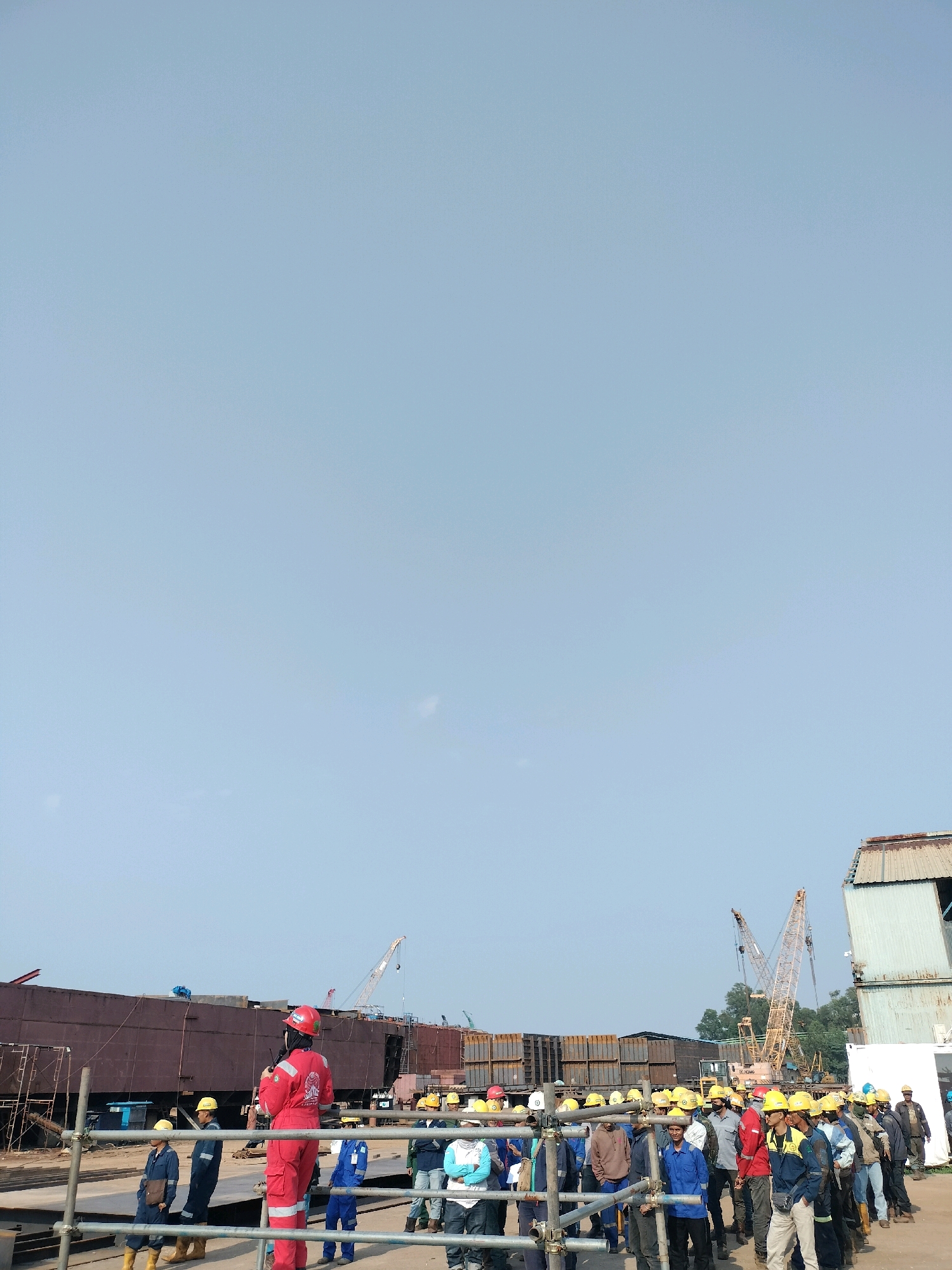 The image depicts a group of workers in a shipyard in Batam, Indonesia. The workers are wearing hard hats and some are in brightly colored jumpsuits, suggesting a construction or industrial setting. They appear to be gathered together, possibly for a briefing or a tour, with one person in a red jumpsuit standing out and seemingly addressing the group. In the background, large structures that could be parts of ships or industrial buildings are visible, along with cranes, indicating active construction or manufacturing. The environment is outdoors, under a clear sky.