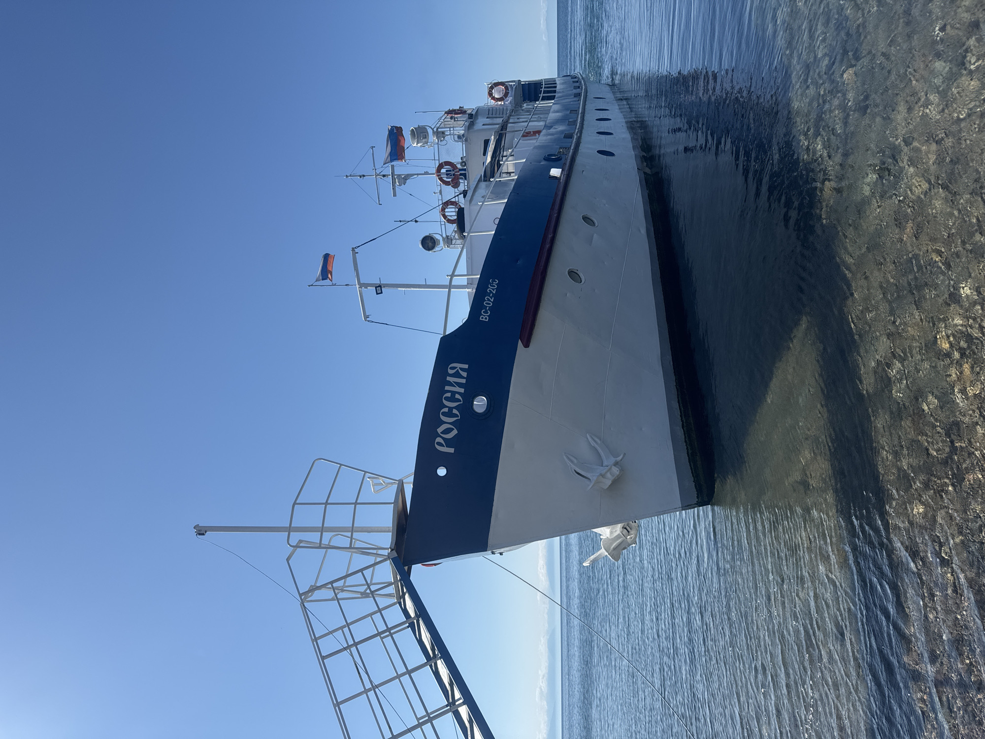 The image shows a white and blue ship named "РОССИЯ" docked in the clear, shallow waters of Lake Baikal near the shore in Listvyanka, Russia. The ship is moored, with its bow facing slightly to the left of the frame. A gangway is extended from the ship to the unseen shore. The water is transparent, revealing the rocky lakebed below. The sky is clear and blue, with a few distant clouds visible on the horizon, where mountains can be seen. The ship has Russian flags flying from its masts. The scene appears calm and serene, suggesting a pleasant day.