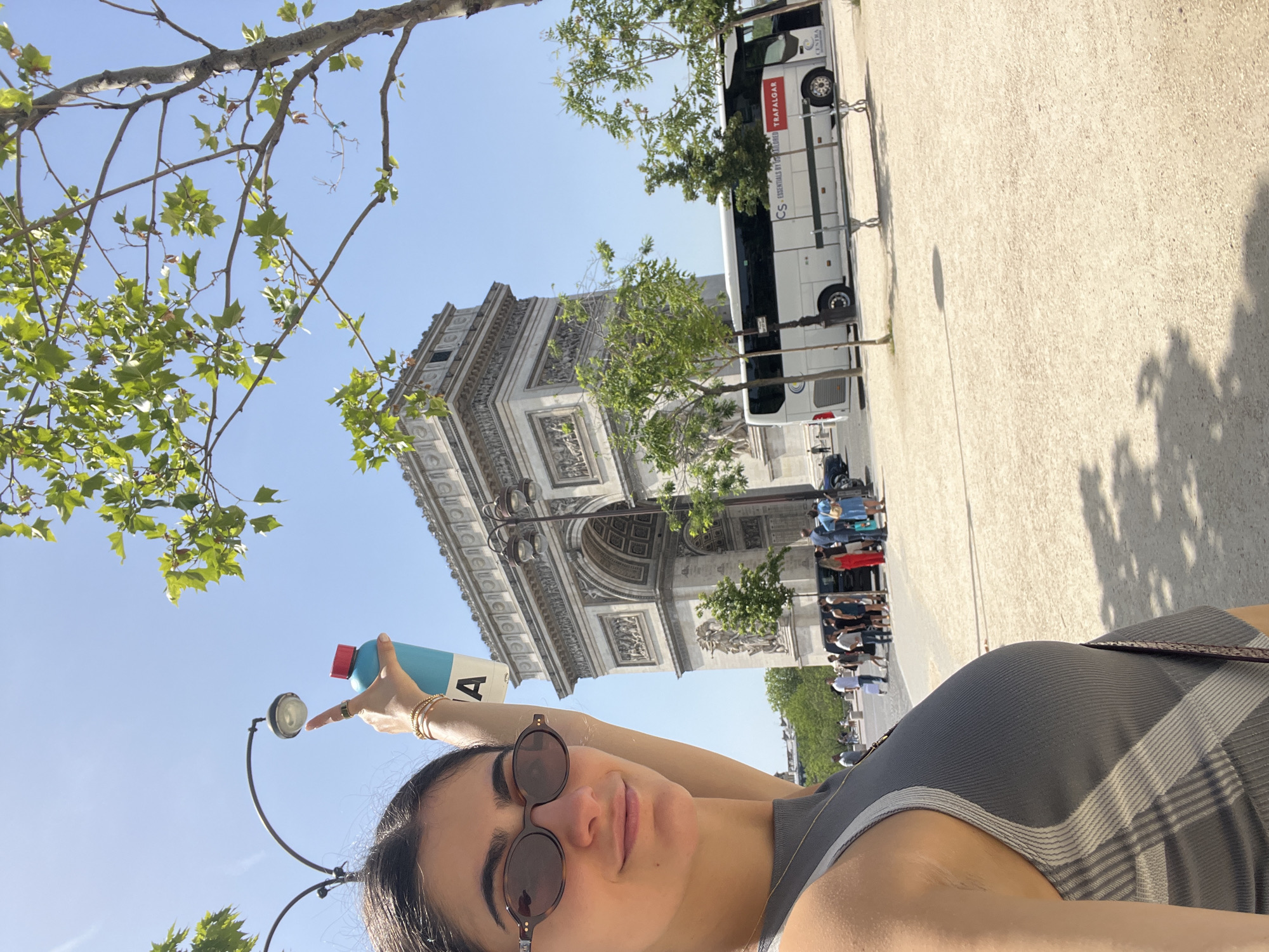 The image shows a woman wearing sunglasses and a gray tank top, holding a blue water bottle with a red lid, and standing in front of the Arc de Triomphe in Paris, France. A white bus is parked nearby. People can be seen in the background. Trees frame the top of the image.