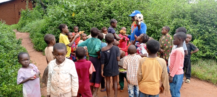 The scene depicts a group of approximately 25 individuals, predominantly children, gathered outdoors on a dirt path in Ngalula, Democratic Republic of the Congo. The environment consists of dense green foliage bordering the path, with a reddish-brown brick or mud-brick structure visible in the background to the left.
Central to the group is one adult, identifiable by a blue long-sleeved shirt, a blue baseball cap with a white logo, and a white head covering underneath. This adult holds a black and white soccer ball in their left hand and a smaller, textured brown sphere in their right hand, appearing to engage with the surrounding children.
The children, ranging in age from toddlers to pre-teens, are dressed in varied, casual attire, some appearing worn. Many children face the adult, observing the objects. Notable actions include a young child on the far left, in a light purple garment, holding a small red object, and two older children in the immediate foreground facing the viewer. The overall disposition of the group suggests an organized activity or presentation by the adult.