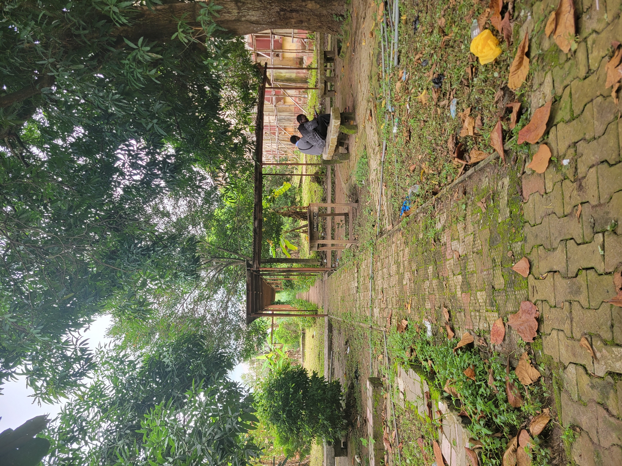 The image depicts an outdoor scene at the University of Uyo, Nigeria, with a pathway leading through a garden area. The pathway is paved with interlocking bricks and bordered by grass and fallen leaves. To the right of the path, a covered seating area with stone benches is visible, where two individuals are seated, appearing to interact with a mobile device. A large tree with dense foliage is prominent on the right side of the image. The background reveals a building under construction or renovation, with scaffolding. The environment appears somewhat neglected, with scattered litter on the ground, including plastic bottles and a yellow plastic bag. Greenery, including bushes and trees, dominates the landscape, suggesting a tropical or subtropical climate.