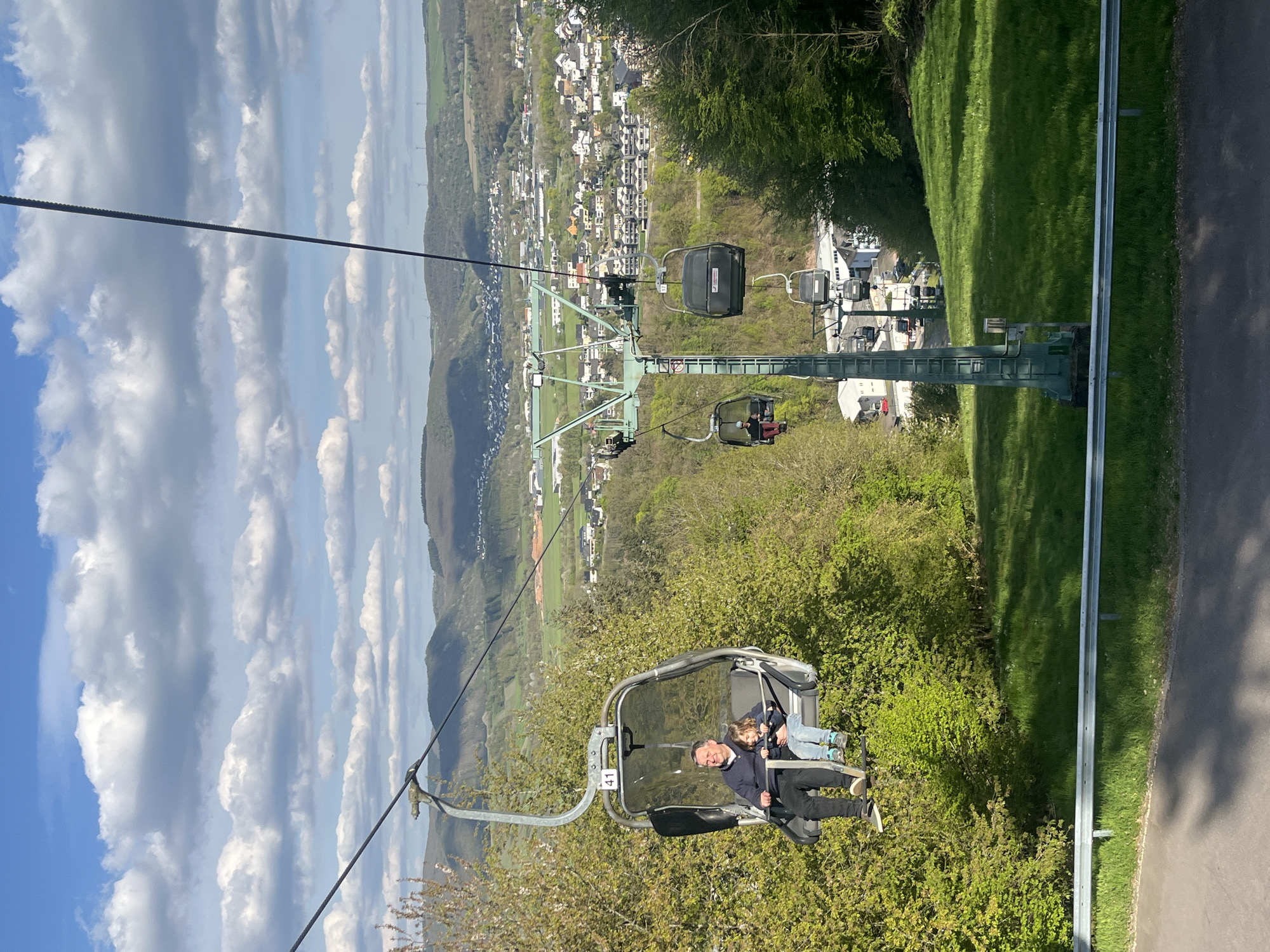 The image shows a view from a chairlift in Saarburg, Germany. A man and a child are riding in one of the chairlift cabins. The chairlift is moving over a green, wooded area and a cityscape. In the distance are hills and a blue sky with clouds. The chairlift structure is visible with other cabins. A paved road with a guardrail is visible below the chairlift.
