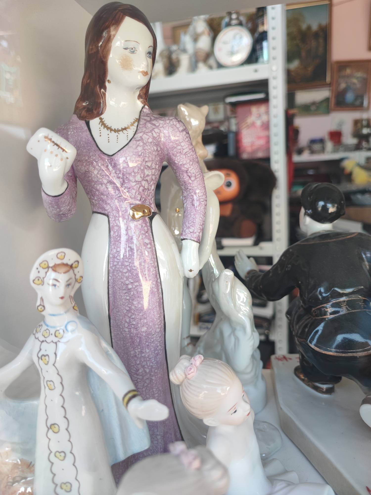 The image shows a collection of porcelain figurines displayed on shelves, likely in a shop or antique store in Pyatigorsk, Russia. In the foreground, a tall female figurine with brown hair, wearing a purple patterned dress, holds a white object. To her left is a smaller figurine of a woman in a white dress with yellow embellishments. To the right of the tall figurine, a boy in a black outfit is crouched down, with his hands extended. Behind them, other figurines and objects are visible, including what appears to be a stuffed toy with large orange eyes on a shelf in the background. The shelving unit is white metal, and behind it, other shelves hold various items, including framed pictures and decorative plates. The lighting suggests an indoor setting.