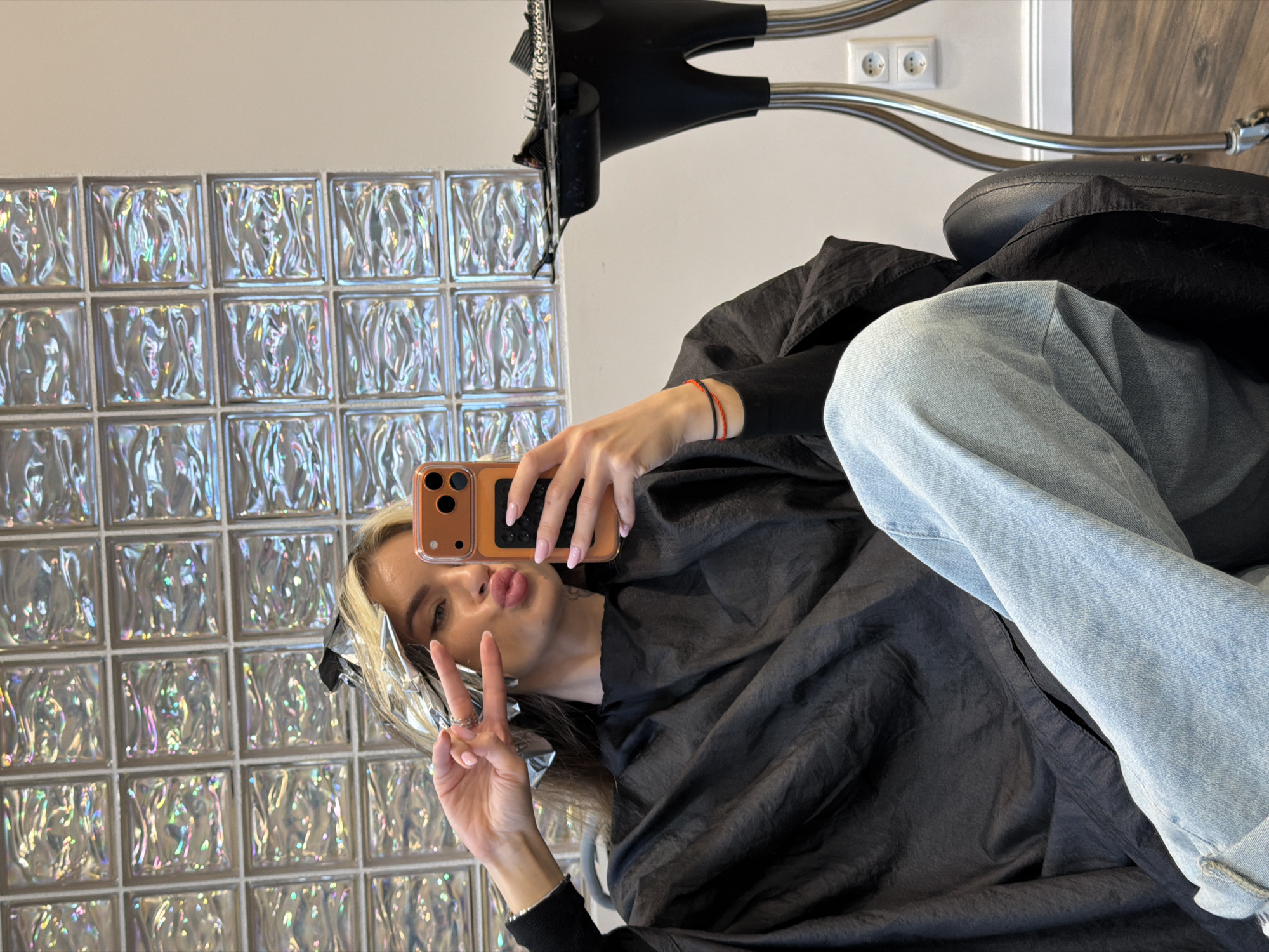 The image depicts a young woman taking a selfie in what appears to be a hair salon. She is wearing light blue jeans and a black hair salon gown. She is holding up a peace sign with her left hand and pouting her lips. Her hair is partially covered with aluminum foil, suggesting she is undergoing a hair coloring process. In the background, there is a wall made of clear glass bricks. To the right, a black salon chair and hair dryer are visible. A white wall with electrical outlets is behind the chair. There is no contextual information to ascertain whether the photograph was taken in Berlin, Germany.