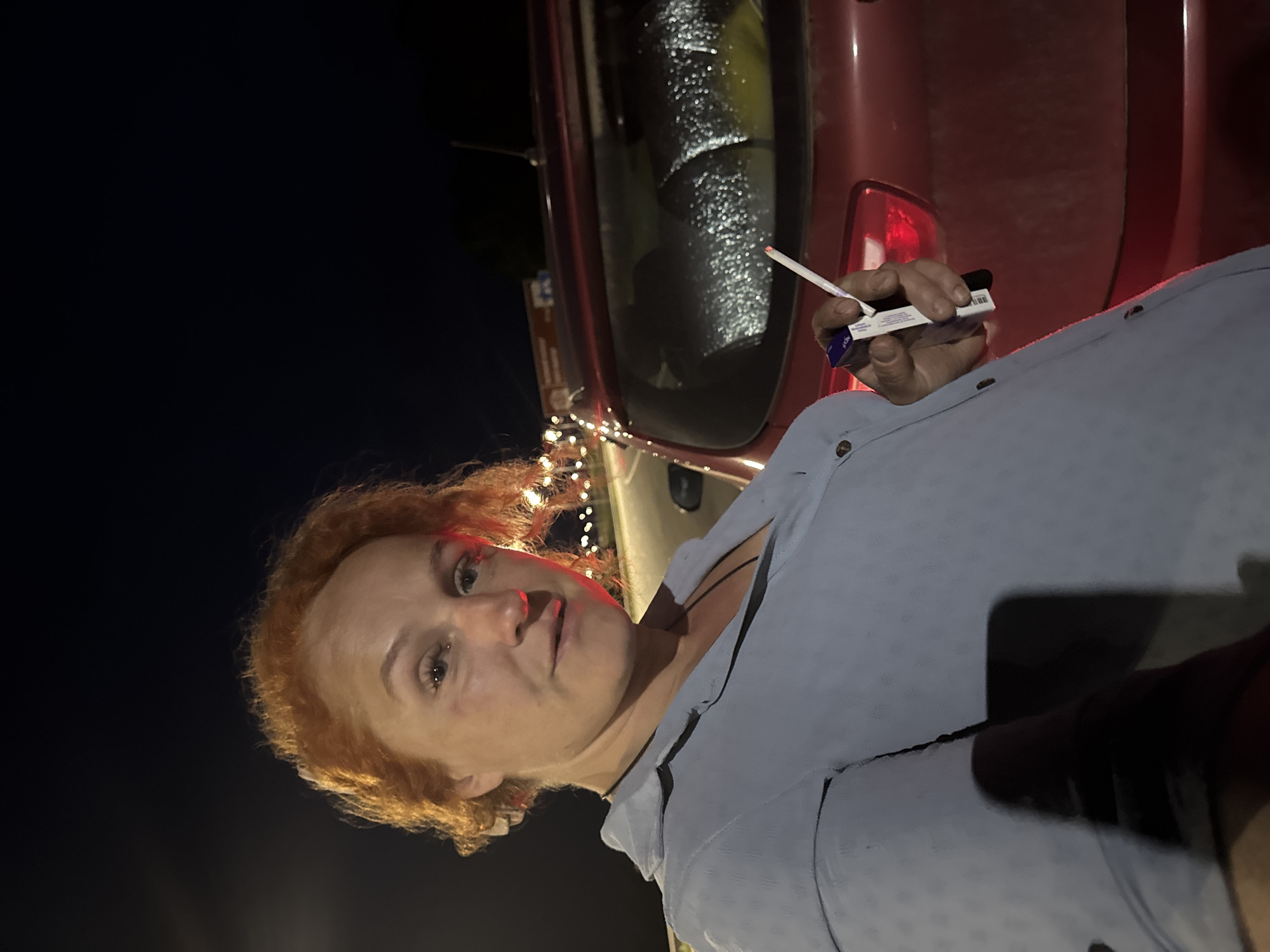 A medium shot captures a woman with auburn hair and fair skin standing at night. She is wearing a light blue button-up shirt and is holding a cigarette in her right hand, which is lit. Next to her is a red car, its back window partially obscured by reflective material. The background is dark, with distant lights suggesting an urban or roadside environment. The woman's gaze is directed towards the camera, and she appears to be looking slightly to the left.