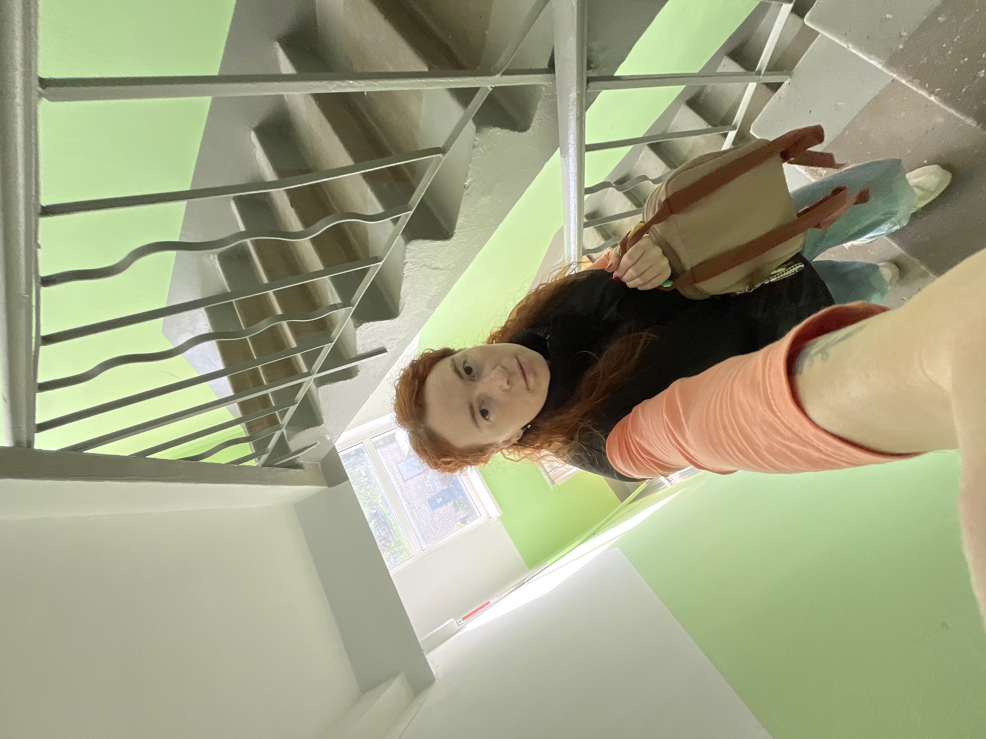 A red-haired woman stands on a staircase in Chaikovsky, Russia. The staircase has a grey metal railing and the walls are painted white and light green. The woman is wearing a black jacket and a peach-colored shirt. She has a beige backpack on her back. A window is visible in the background.