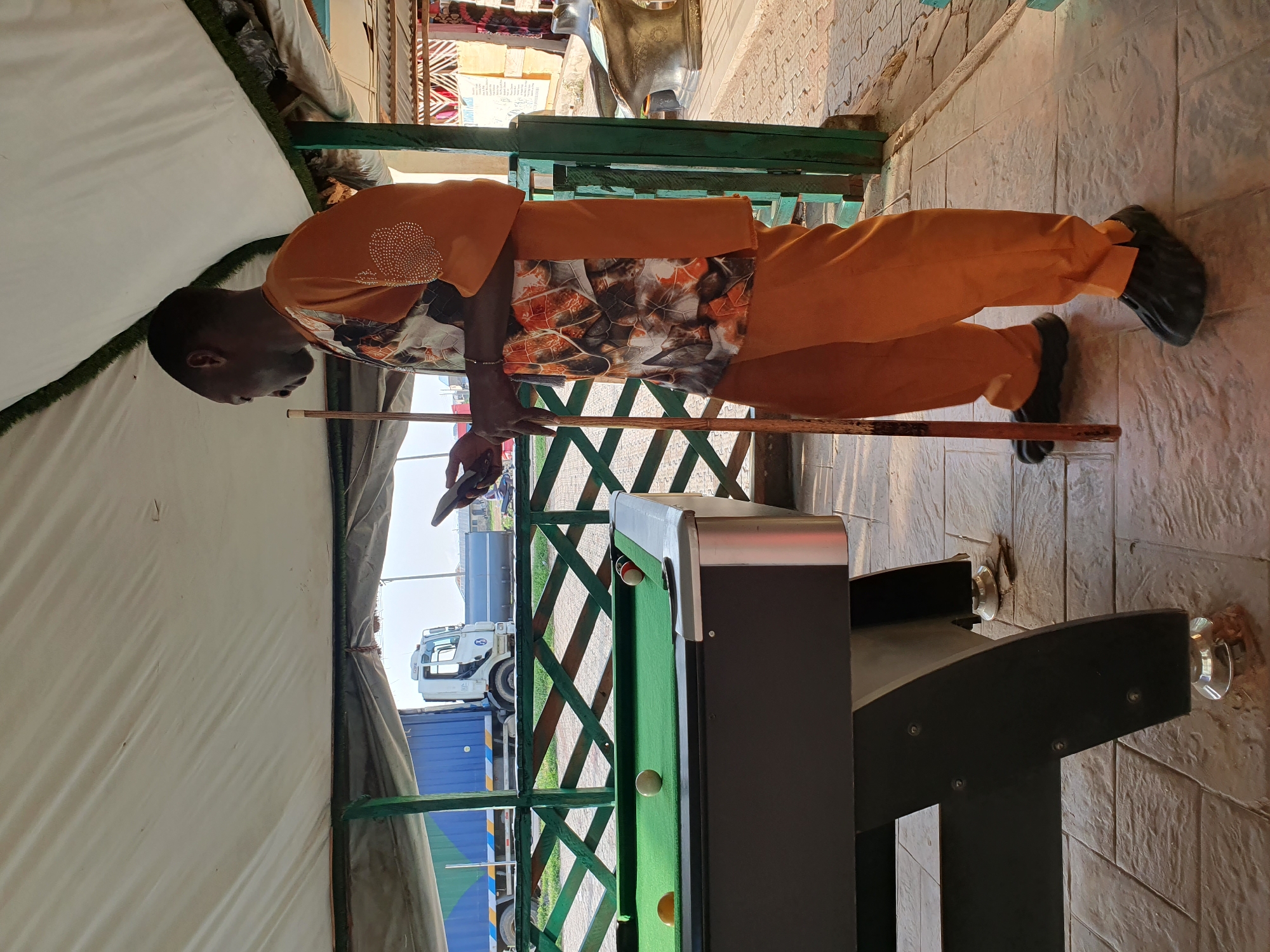 A man is standing next to a pool table in Lokoja, Nigeria. The man is wearing an orange shirt with a leaf print and orange pants. He is holding a pool cue in his right hand and a smartphone in his left hand. There are several billiard balls on the pool table. The pool table is green and is set up outdoors under a white canopy. In the background, a truck and trailer are visible. The ground is paved with stone tiles.