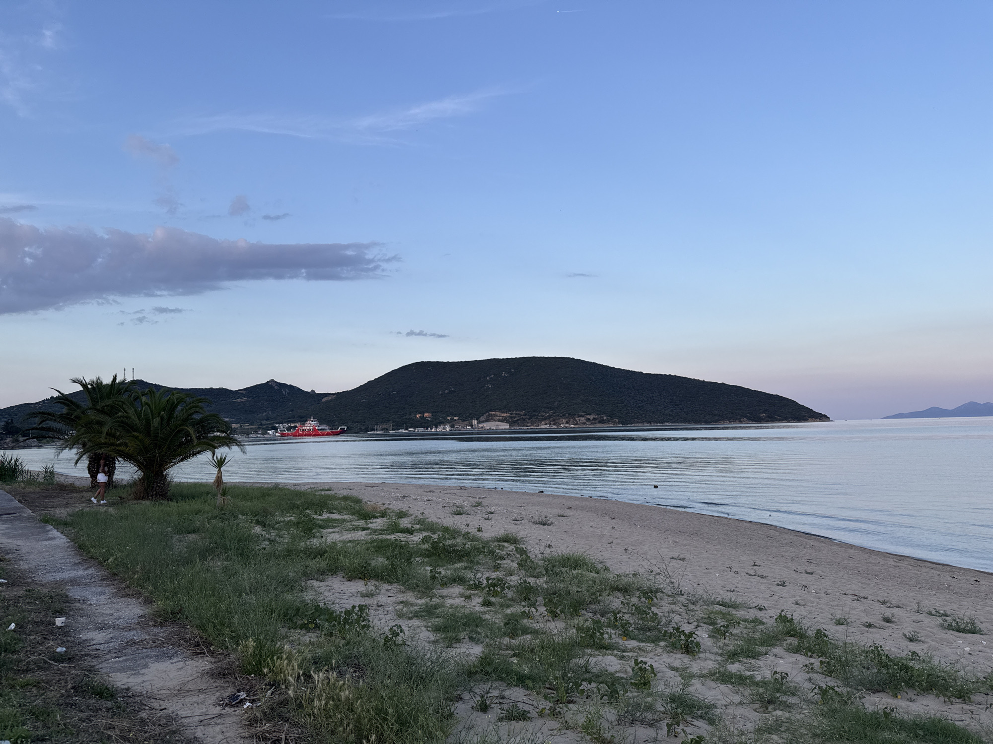 A wide shot captures a serene coastal scene at dusk in Nea Peramos, Greece. The sky is a gradient of pale blue to light purple, with a few wispy clouds scattered across the horizon. A large, dark green, treed hill dominates the background, sloping down to the tranquil, pale blue water. A red ferry boat is visible on the water in the distance, near the shore. The foreground features a sandy beach with sparse patches of green grass and some scattered debris. To the left, a paved pathway runs alongside the beach, leading towards a prominent palm tree. A person in light-colored clothing stands near the palm tree, facing away from the camera. The overall atmosphere is peaceful and quiet, indicative of the twilight hour.