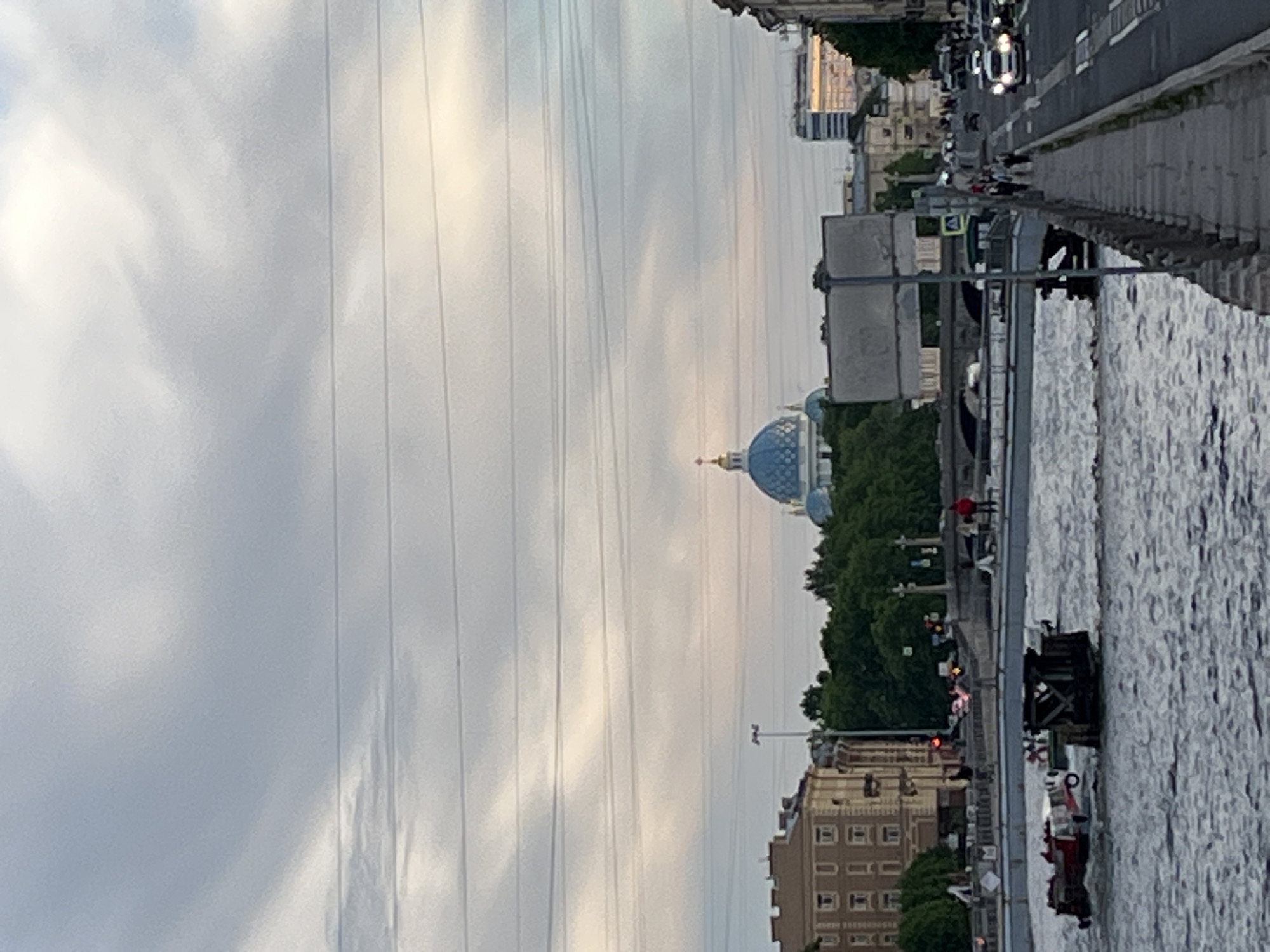 The image depicts a panoramic view of a cityscape in Saint Petersburg, Russia, featuring a wide body of water, a busy street, and architectural landmarks under an overcast sky. The scene, if viewed with correct orientation (rotated 90 degrees counter-clockwise), shows a river or canal occupying the right side of the frame, its surface rippled by current or movement, with a few small boats, possibly tourist vessels, present.
Parallel to the water runs a paved promenade, where several indistinct human figures can be seen walking. To the left of the promenade, a multi-lane street carries vehicular traffic, including cars, one of which has illuminated headlights, suggesting either dim light conditions or late afternoon/early evening. Various buildings of differing heights and architectural styles line the street, partially obscured by green trees.
In the middle distance, a distinctive blue-domed cathedral topped with a golden spire stands out against the horizon, strongly resembling the Trinity Cathedral (Troitsky Sobor). Numerous thin, parallel overhead wires stretch across the expansive, cloudy sky. The overall atmosphere is one of routine urban activity. No discernible text is visible in the image.
