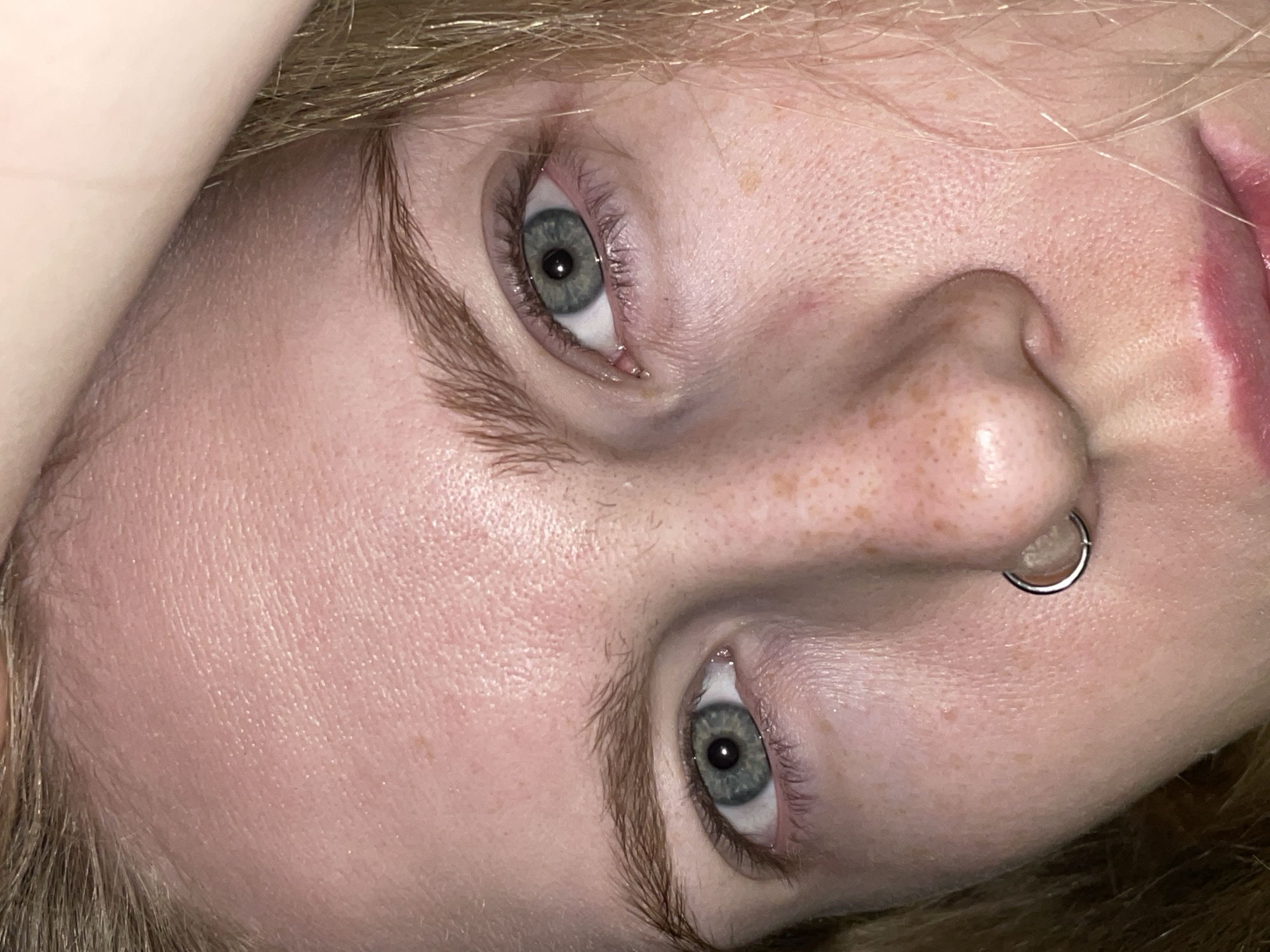 A close-up, angled shot captures the face of a person, likely lying down, with light blonde or strawberry blonde hair framing their features. The perspective is tilted, showing the person's head rotated counter-clockwise.
The subject has striking light blue or grey-green eyes that are open and directly facing the viewer. There is a noticeable redness around the eyes, particularly on the lower lids, which could suggest irritation or fatigue. Long eyelashes are visible. The skin tone is fair, with a scattering of prominent freckles across the nose and cheeks. Pores are visible on the skin, indicating a high-resolution image.
A shiny, silver-colored circular septum piercing is visible through the nose, adding a distinctive detail. Only the far right corner of the person's mouth and part of the upper lip are visible, tinted a natural pink or with a hint of lip color. The hair is light, appearing fine and slightly messy, contributing to the casual nature of the shot.
The lighting appears to be indoor or artificial, creating soft highlights on the forehead and nose. No specific activity beyond looking at the camera is depicted, and the expression is relatively neutral, perhaps thoughtful or tired. There is no visible text in the image. The location context provided is Izhevsk, Russia.
