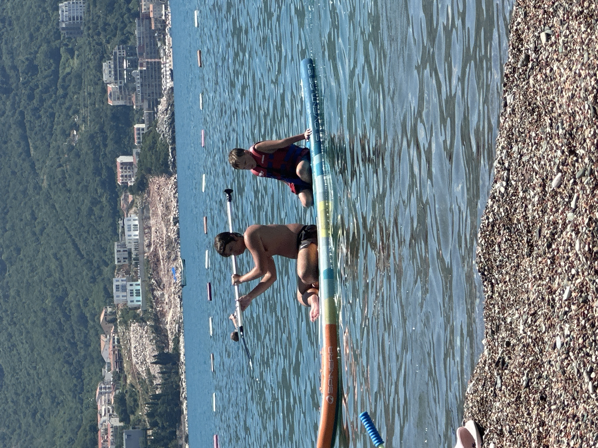 Two young males are on a stand-up paddleboard in the sea. One, wearing a red and blue life vest and dark shorts, is kneeling and holding onto the board. The other, shirtless and wearing dark shorts, is also kneeling on the board and holding a paddle. The paddleboard is colorful with the text "SPINERA" visible on it. The water is a clear blue with ripples. In the background, there is a coastline with buildings and lush green hills. The sun appears to be shining, indicating a daytime activity. The scene is set in Boreti, Montenegro, a coastal town known for its beaches and mountainous backdrop.