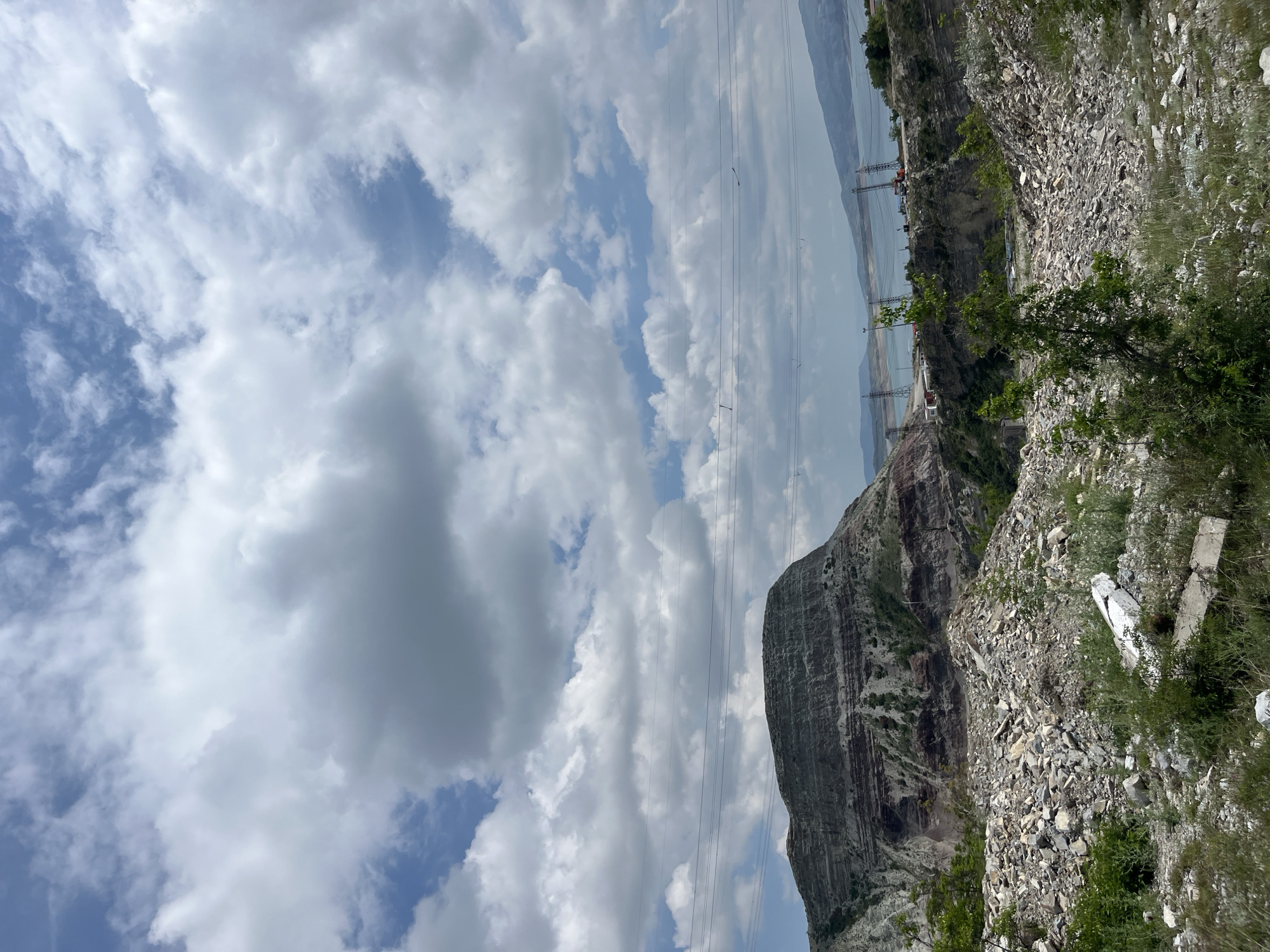 The image depicts a mountainous landscape under a partly cloudy sky in Chirkey, Russia. A large, striated rock formation dominates the foreground, with power lines stretching across the frame towards what appears to be a lake or reservoir in the background. The foreground also includes rocky terrain with sparse vegetation. In the distance, a structure resembling a bridge or dam can be discerned. No people are visible in the image.