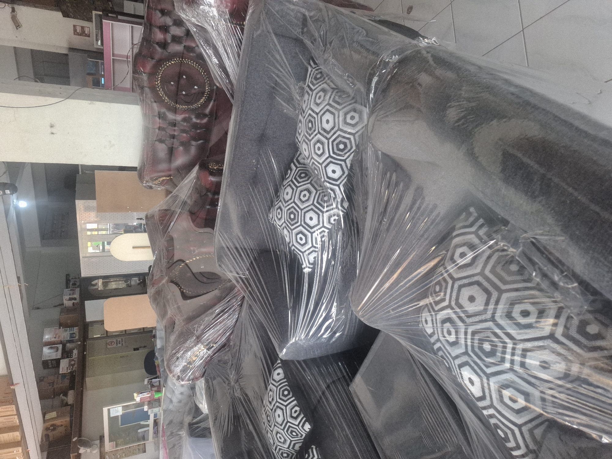 This image depicts a furniture store located in Banjarmadu, Indonesia. The scene is filled with various upholstered furniture items, primarily sofas, all wrapped in clear plastic for protection. In the foreground, a dark gray sofa with geometric-patterned throw pillows is visible, also covered in plastic. Behind this, other sofas and seating arrangements can be seen, including a dark red tufted sofa with ornate gold detailing. The store appears to be an indoor space with a concrete floor and white walls. Shelving units holding boxes and various items are visible in the background, suggesting a retail or storage environment. The lighting is somewhat dim, with artificial lights visible on the ceiling. There are no people visible in the image.