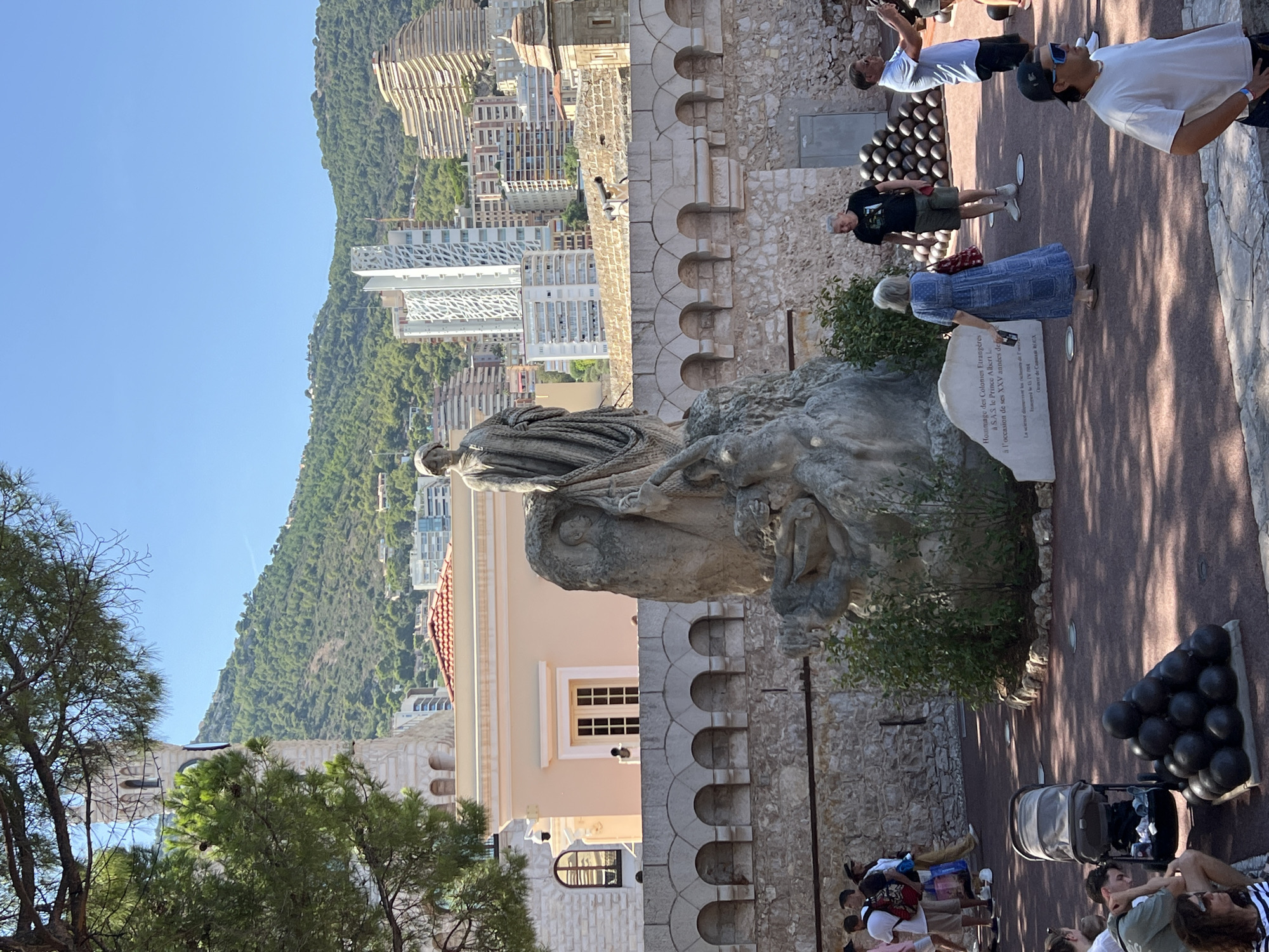 The image depicts a sun-drenched outdoor plaza in Monaco, likely within the Prince's Palace area, given the presence of cannons and a significant monument. In the foreground, a group of tourists are visible. To the left, several individuals stand near a stroller, while a pyramid of dark, spherical cannonballs rests on the ground. A statue, carved from light-colored stone, dominates the center of the frame. It features a draped figure standing above a sculpted mythological creature. To the right of the statue, a woman in a blue dress is walking away from the camera, holding a red bag. A man in a black t-shirt and shorts stands nearby, and another man in a blue t-shirt is taking a photo with his phone.
Behind the plaza, a stone wall with arched openings forms a terrace. Beyond the wall, several buildings are visible, including a tall, white, modern skyscraper and older, terraced structures. A lush, green, mountainous landscape forms the backdrop under a clear blue sky. A section of a stone tower, possibly part of the palace complex, is visible on the far left, partially obscured by a pine tree. A plaque at the base of the monument contains French text, suggesting it is a dedication.