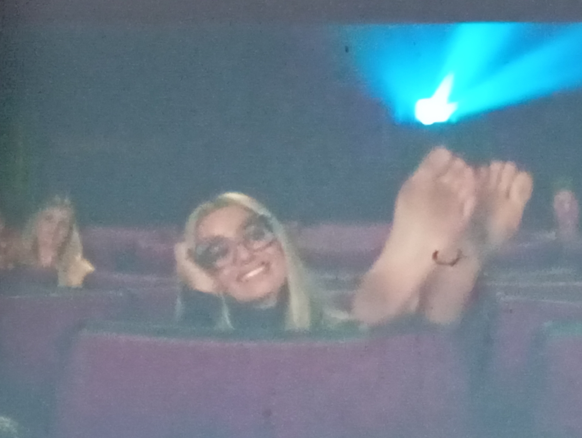 A group of people are seated in what appears to be a theater or cinema, watching a bright blue light projected onto a screen. The focus is on a woman in the foreground who is reclining in her seat with her feet up, seemingly relaxed and enjoying the show. She has blonde hair and is wearing glasses and a dark top. Her bare feet are clearly visible, with the toes spread. Another woman with blonde hair is visible in the background to the left, also appearing to be watching the projection. The overall mood seems casual and leisurely, suggesting a movie viewing or a presentation. The lighting indicates it's likely nighttime or an indoor venue. The context of Sofiivska Borshchahivka, Ukraine, suggests this is a local event.