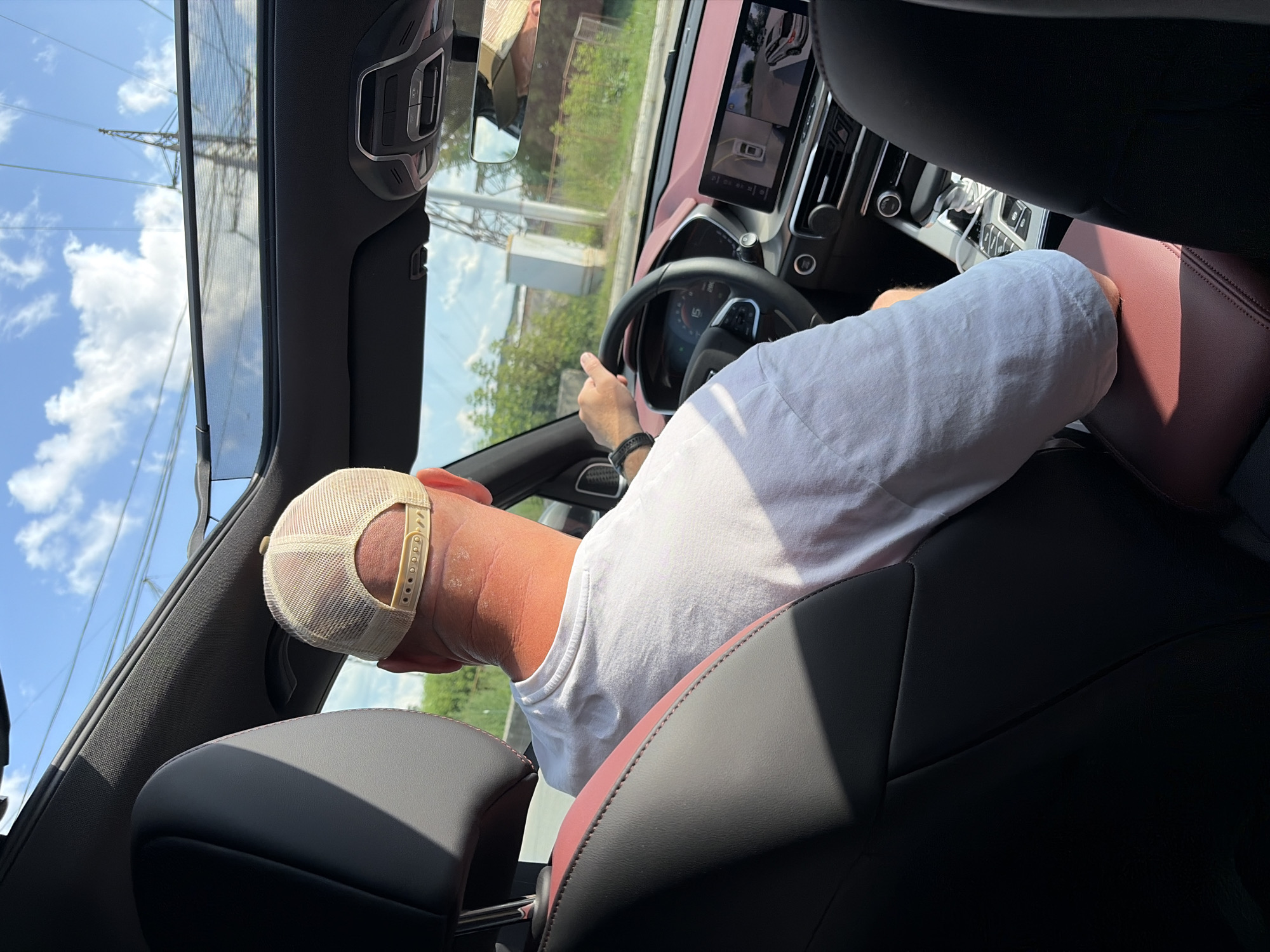 The image shows a man driving a car on a street in Chaikovsky, Russia. He is wearing a white t-shirt and a tan baseball cap. The car has a sunroof, and the interior appears to be modern with a large touchscreen display showing a 360-degree camera view. The exterior scenery suggests an urban or suburban environment with trees and utility poles visible. The weather is sunny with clouds in the sky.