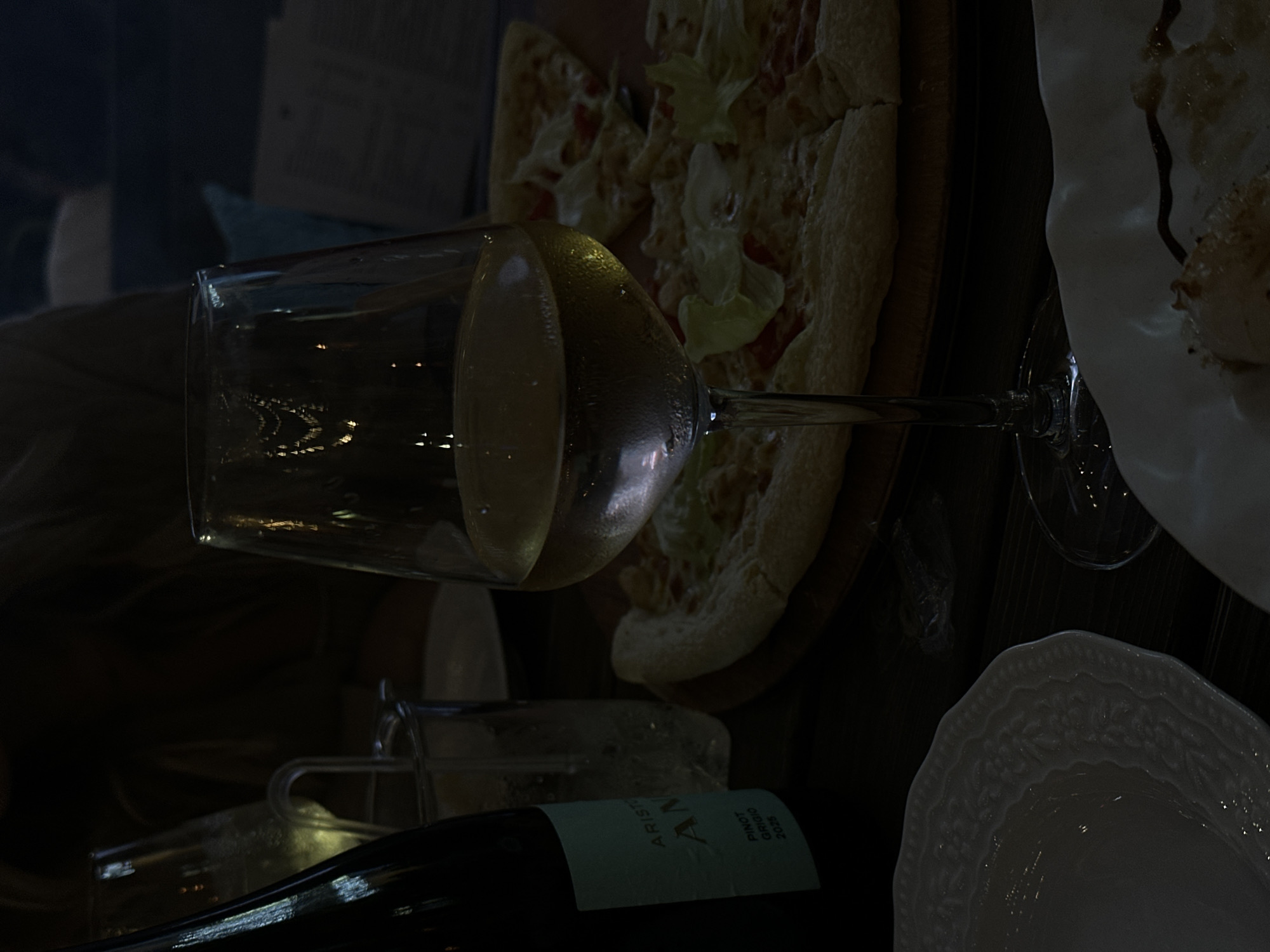 This image depicts a dimly lit dining scene, likely in a restaurant, with a focus on food and drink. A glass of white wine stands prominently in the foreground, condensation visible on its surface, indicating it is chilled. To the right of the wine glass, a pizza is presented on a wooden board. The pizza appears to be cut into slices and topped with cheese and some green leaves, possibly lettuce or herbs. A dark glass bottle, likely containing wine, is positioned to the left of the wine glass. Its label, partially visible, reads "ARISTO" and "AN", with "PINOT GRIGIO 2025" at the bottom. A tall, clear glass filled with ice and liquid is situated behind the wine bottle. Another white plate with a decorative rim is visible in the lower left corner. The overall lighting is low, suggesting an evening meal. There are no visible people or specific landmarks that would indicate a location in Izhevsk, Russia.