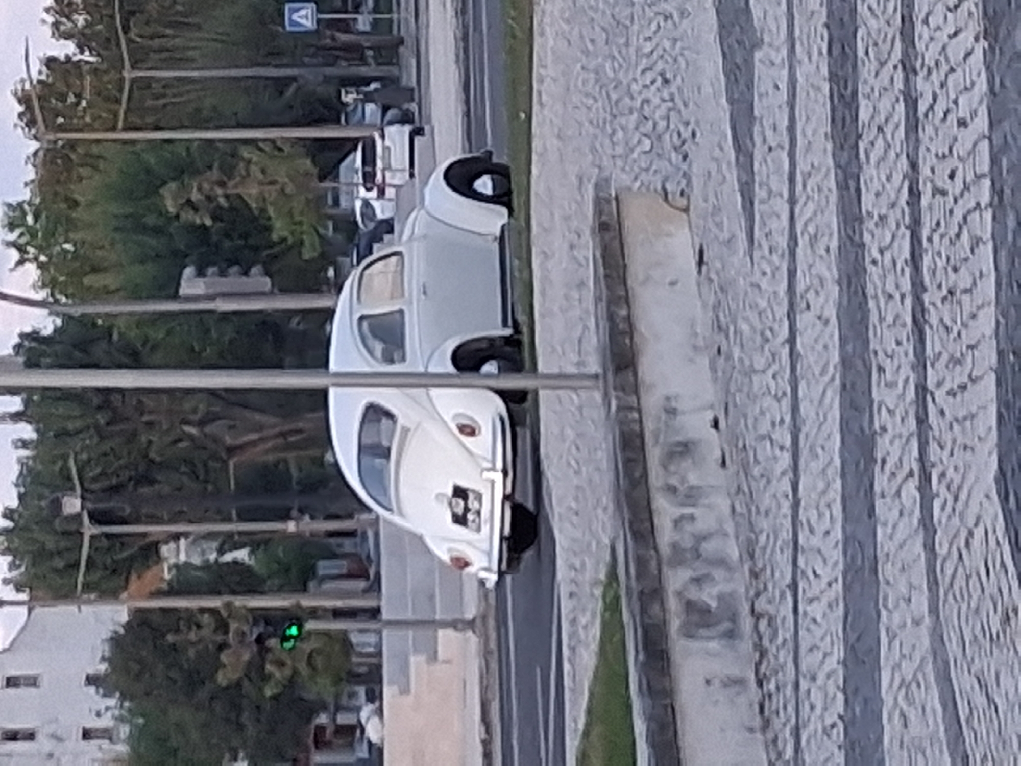A white Volkswagen Beetle is parked on the roadside, adjacent to a sidewalk with a patterned, grey-and-white paving. Beyond the sidewalk, there's a strip of green grass and then the road, with other vehicles parked. Trees and streetlights line the opposite side of the road, with buildings visible in the background. A traffic light showing green is also visible. The scene suggests a street in the city of Figueira da Foz, Portugal.