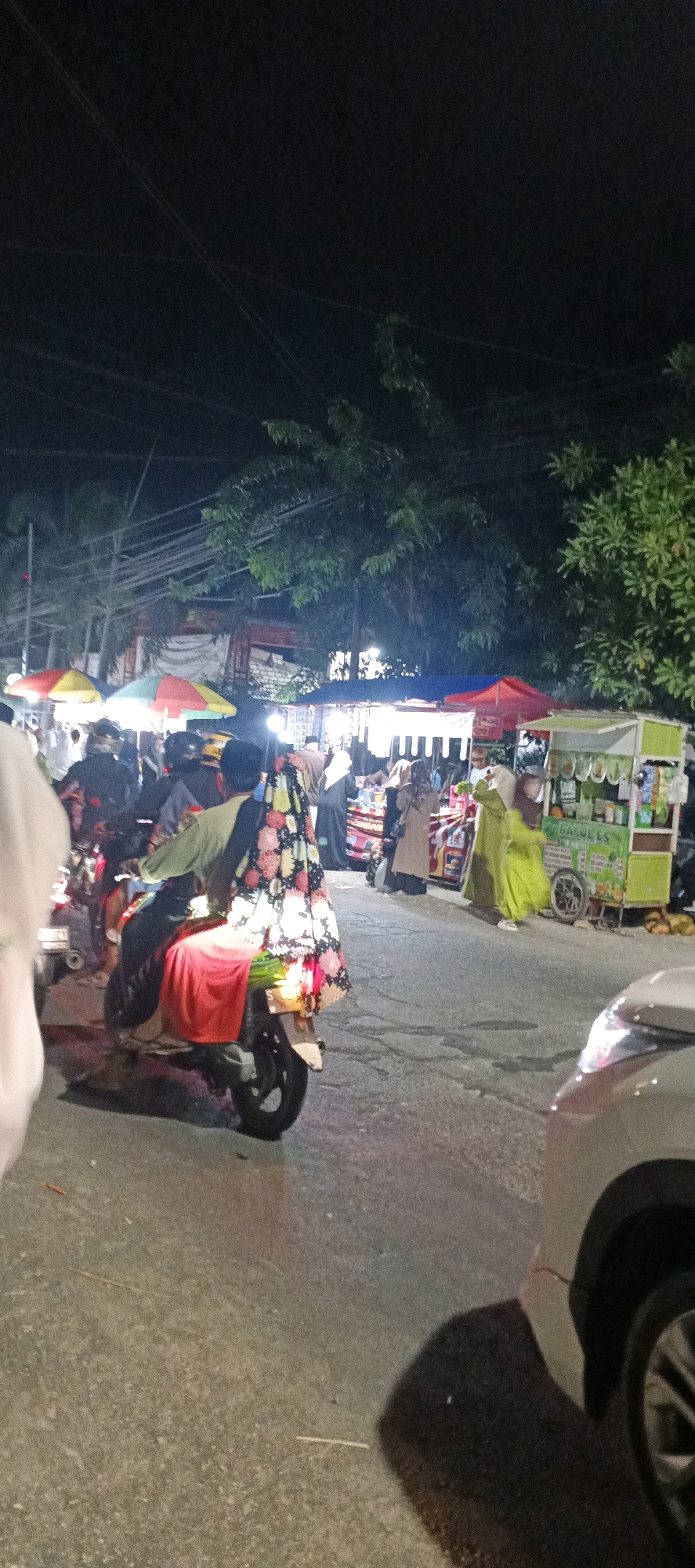 The image depicts a nighttime street scene in Sungelebak, Indonesia, likely a marketplace or a busy thoroughfare with street vendors. In the foreground, a person rides on the back of a motorcycle, draped in a colorful, floral patterned fabric that also covers the rear of the motorcycle. They are wearing a red garment underneath the fabric. To the right, a white car is partially visible.
In the background, several street stalls are set up, illuminated by artificial lights. One stall, on the right side of the image, is a bright green and white structure with text that appears to be a menu or business name, possibly related to food or drinks. Various people are browsing or working at these stalls, dressed in a mix of casual and traditional clothing, many wearing hijabs.
The street is paved, and overhead power lines are visible against the dark night sky, indicating an urban or semi-urban environment. Trees with dense foliage are present behind the stalls. The overall atmosphere suggests a lively, nocturnal community gathering or commercial activity.